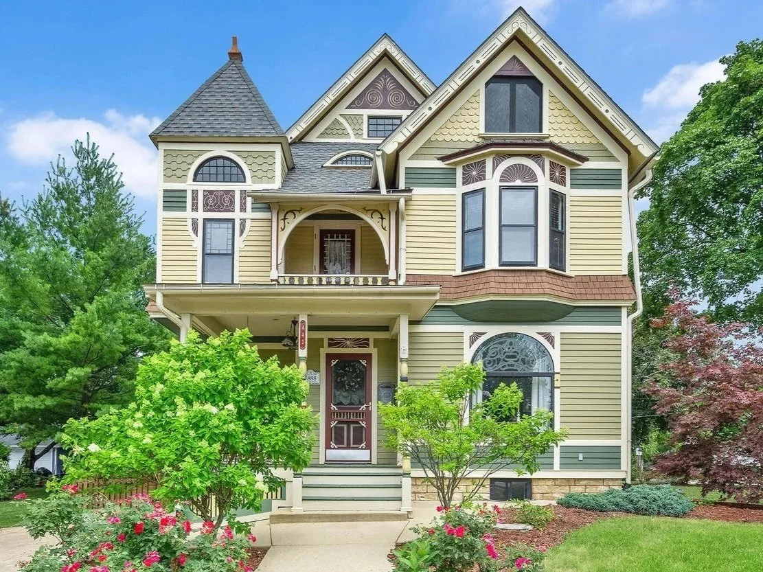 A Victorian-style house with yellow and green siding, a steep gray roof, and a small front porch surrounded by trees and bushes.