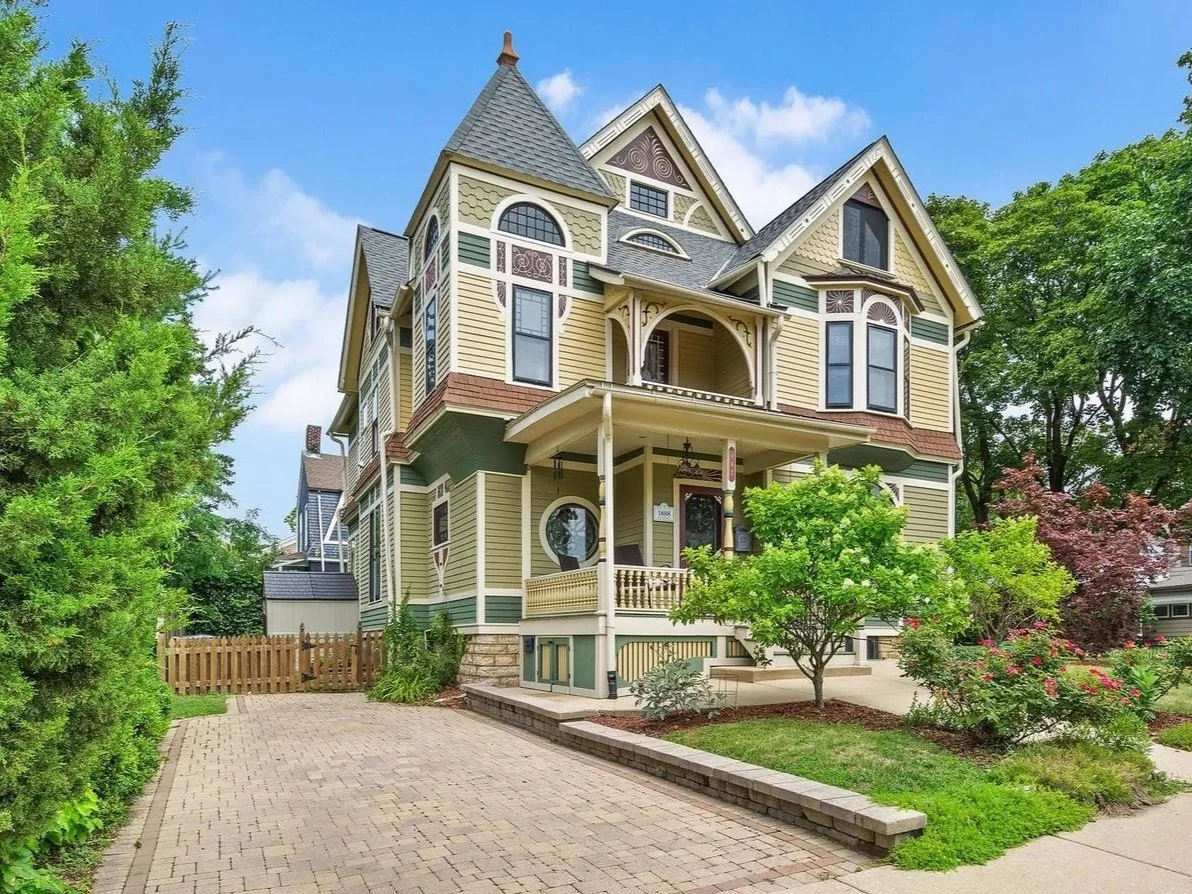 A large Victorian-style house with yellow and green siding, a turret, and a front porch surrounded by lush greenery.