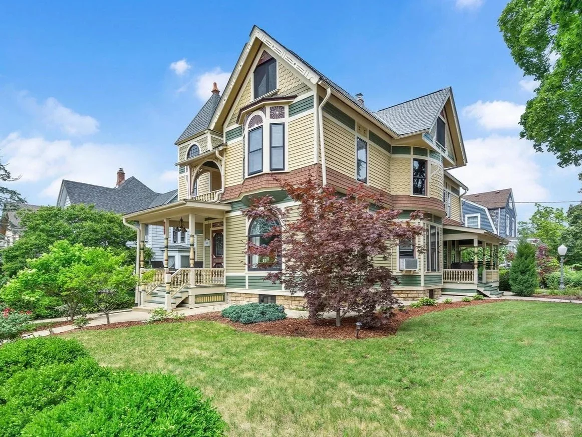 A large Victorian-style house with a bay window, a front porch, and a steep gabled roof, surrounded by a manicured lawn and green trees under a partly cloudy sky.