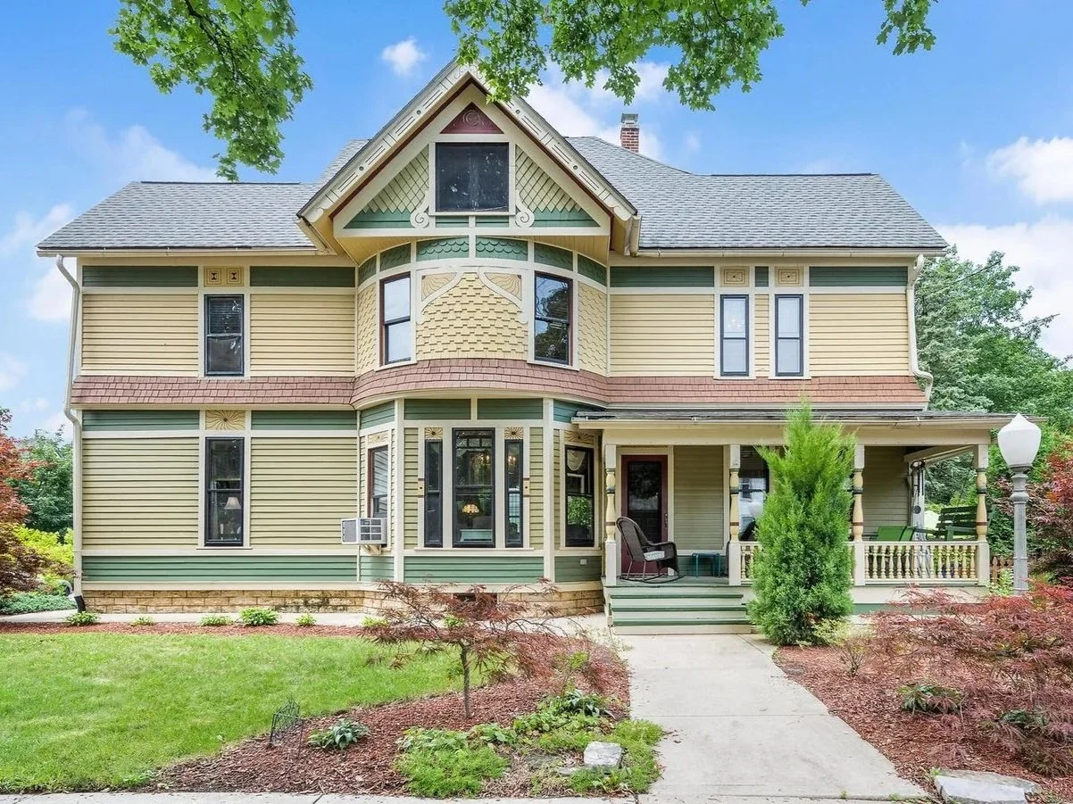 A two-story Victorian-style house with yellow and green exterior, decorative trim, a front porch with chairs, and a sidewalk leading to the entrance, surrounded by landscaped yard with small trees and bushes.