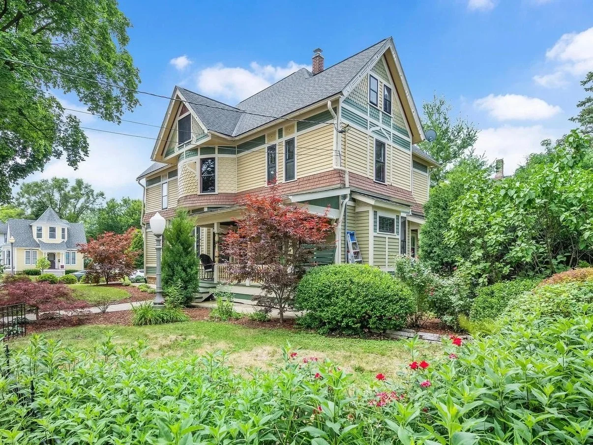 A large Victorian-style house with yellow and green siding, surrounded by a lush garden with trees, shrubs, and flowering plants, under a partly cloudy sky.