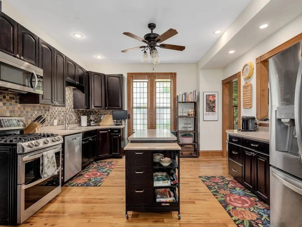 Kitchen with dark cabinets, stainless steel appliances, a small kitchen island, a ceiling fan, colorful floral rug, wooden floors, and a window with wooden blinds.