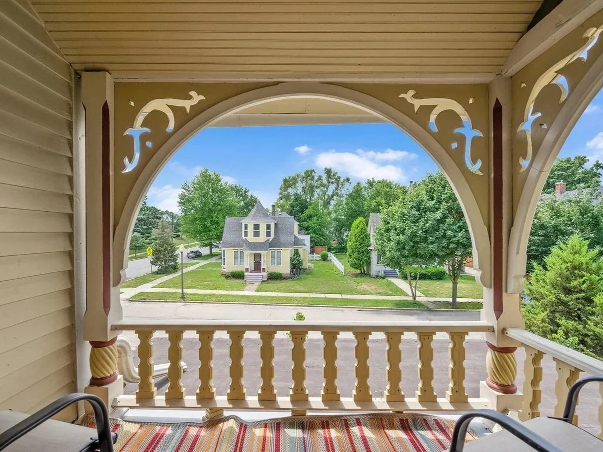 View from a porch with ornate decorative trim, overlooking a residential street with a yellow house, lush green trees, and a sunny sky.