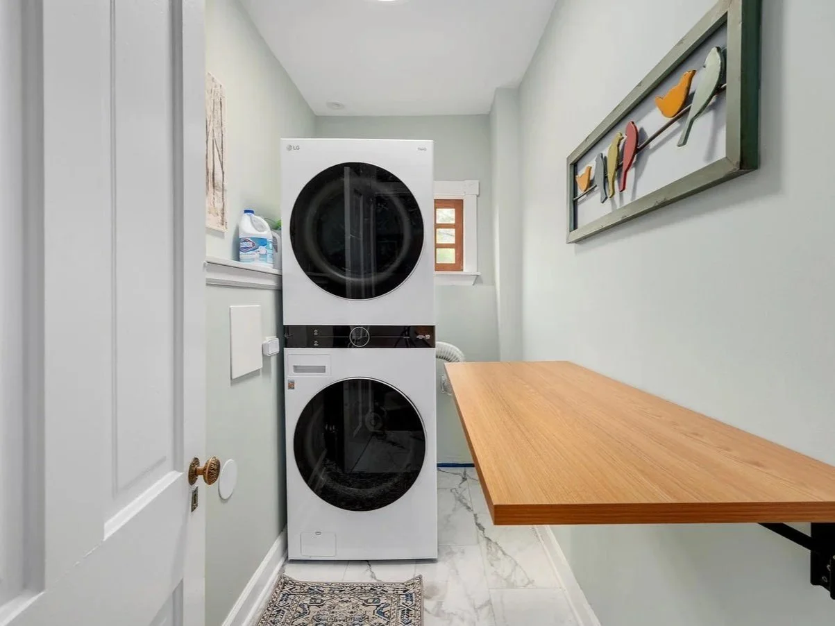 Small laundry room with stacked washer and dryer, a window, an art piece with birds on the wall, a wooden folding table, and cleaning supplies.