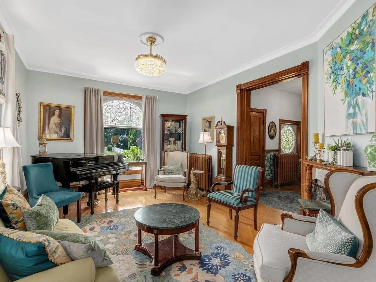 Living room with a grand piano, vintage armchairs, a sofa with pillows, a round coffee table, and decorative items, with a large window and wooden trim.