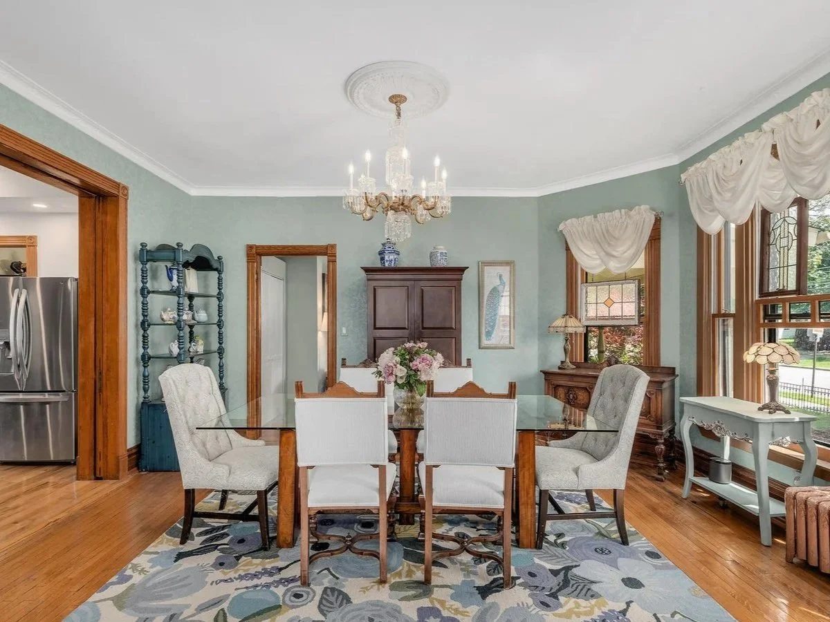 Dining room with a glass-top table, six upholstered chairs, a floral centerpiece, a chandelier, wooden trim, and large windows with curtains.