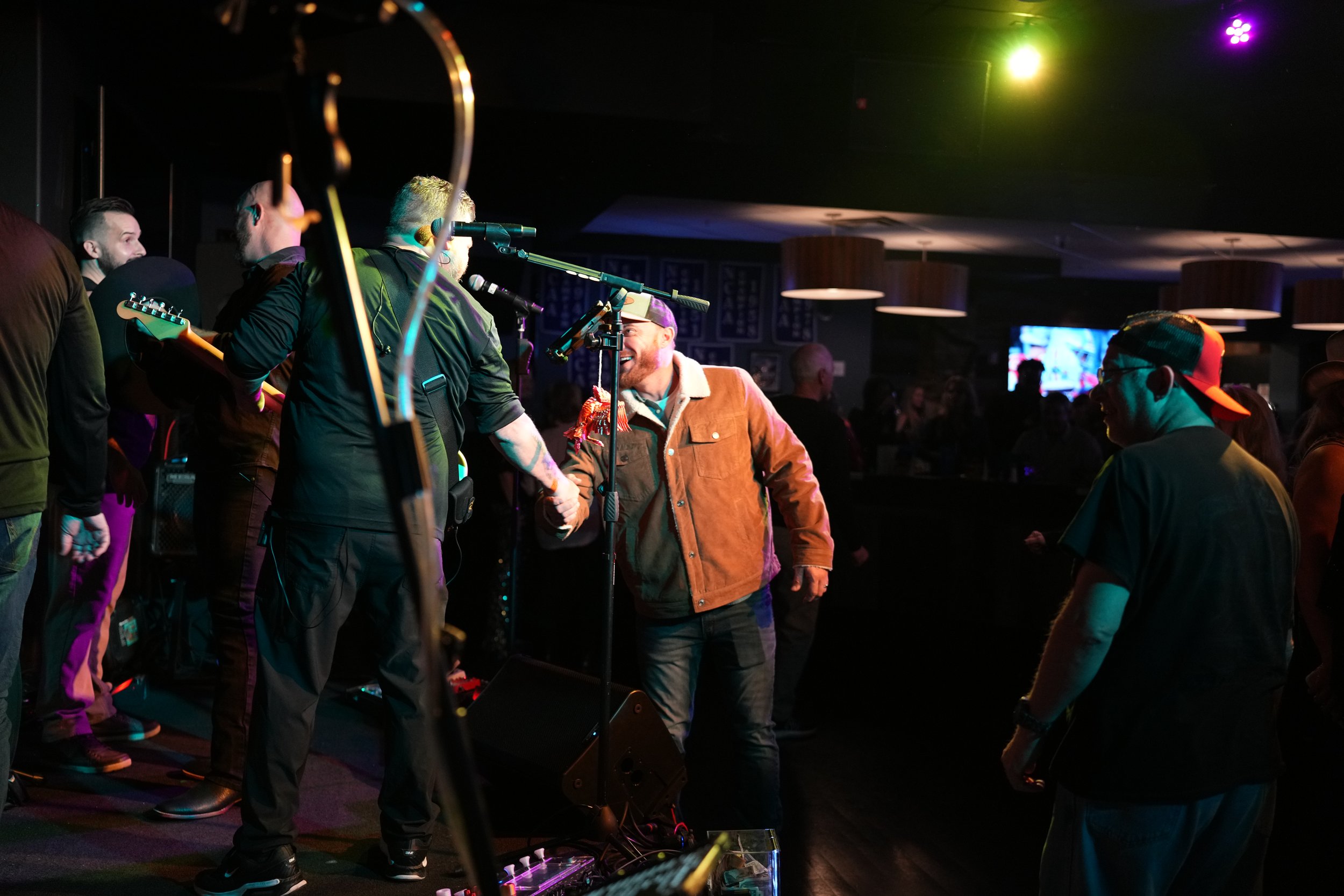 People enjoying live music performance in a dimly lit venue with colorful stage lights, some interacting and smiling.