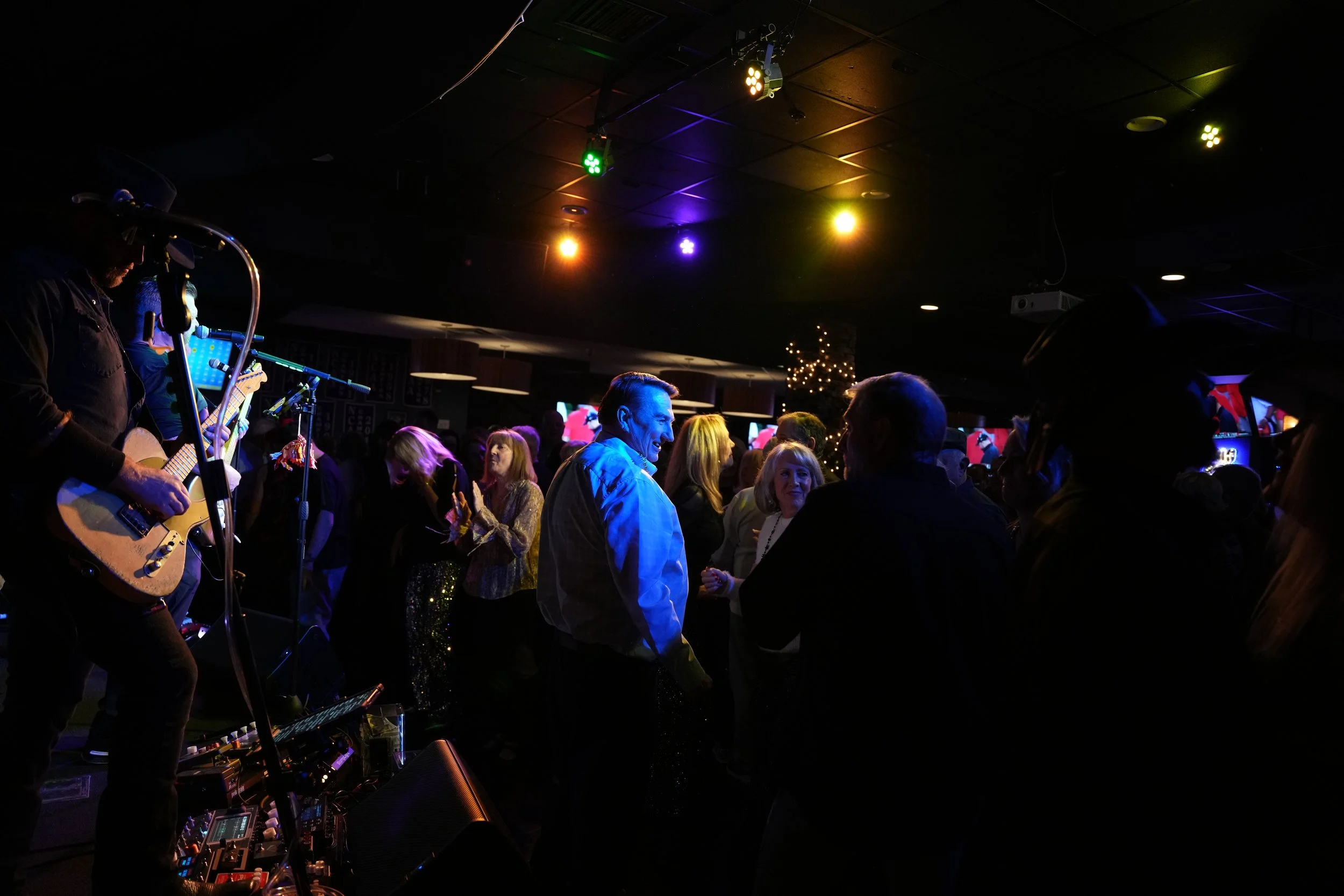 A lively indoor event with a band performing on stage, colorful spotlights overhead, and a crowd dancing and socializing.