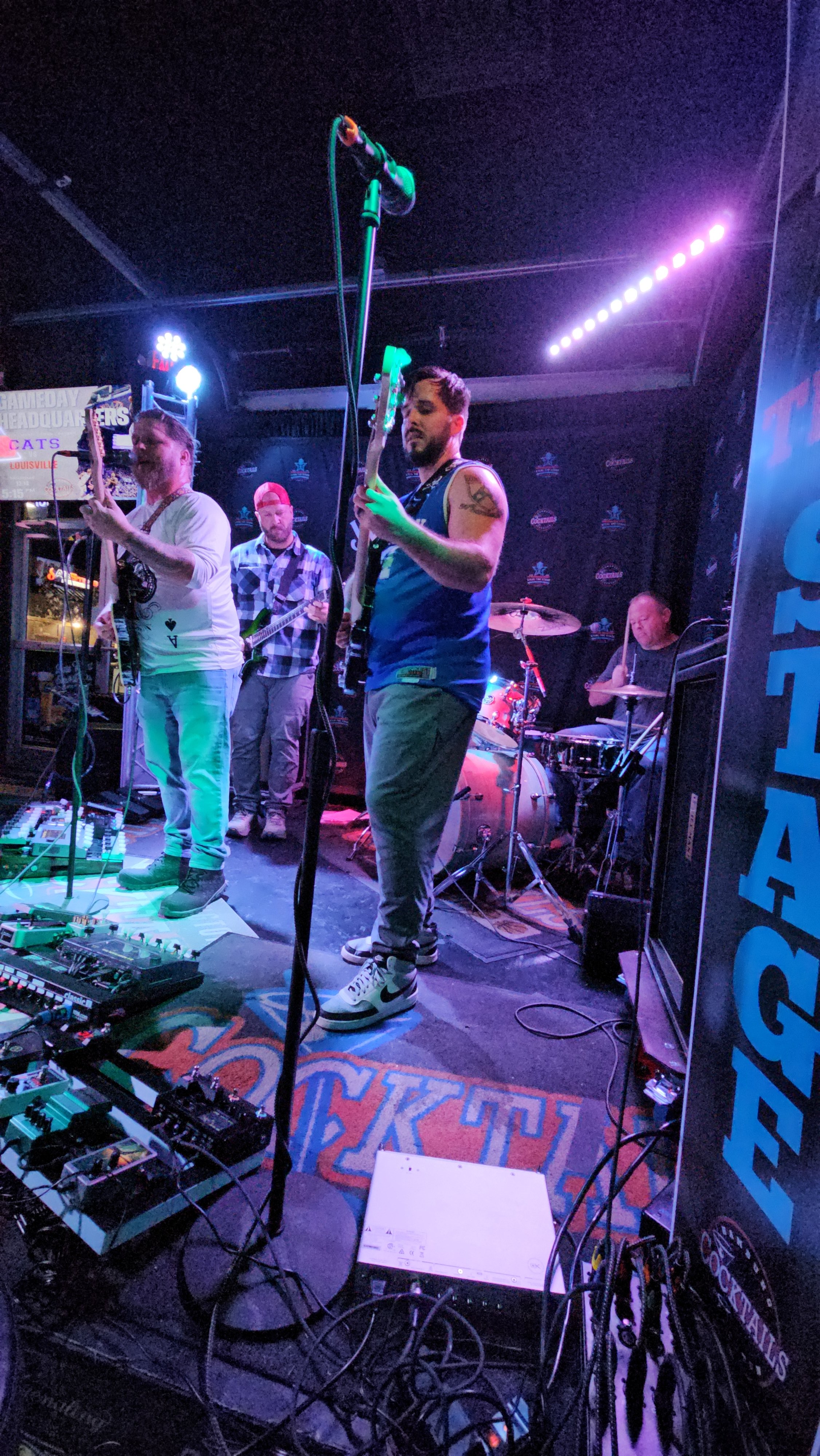 A live band performing on stage at a music venue with colorful lighting, with four musicians playing guitars, drums, and pedals.