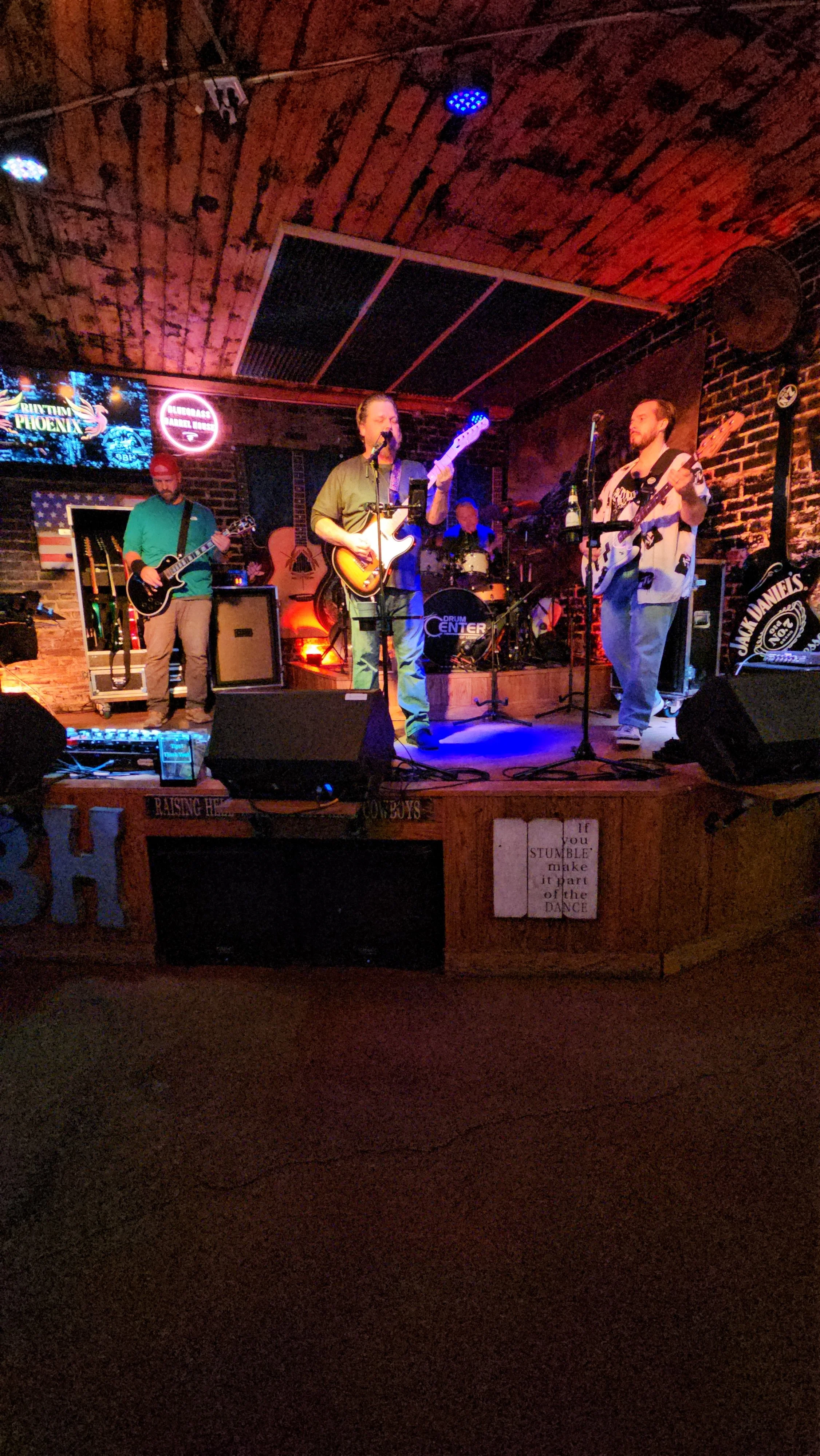 A band performing on a small stage inside a dimly lit bar or club, with brick walls and wooden ceiling. The band includes three male musicians playing guitars and drums, with various equipment around them. Neon signs and a TV screen are visible in th