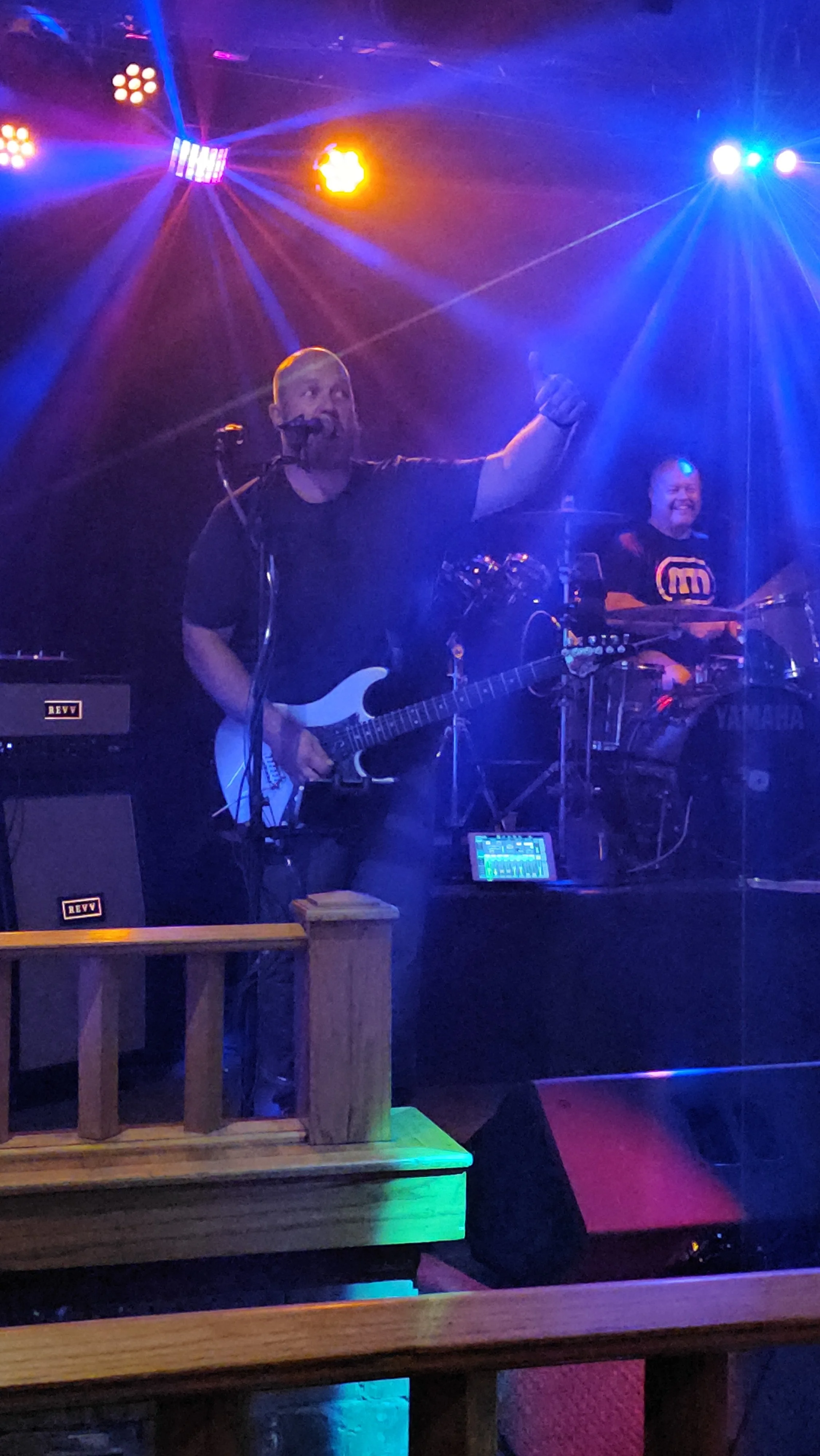 Two musicians performing on stage with colorful lighting and effects, one playing electric guitar and singing into a microphone, the other on drums.