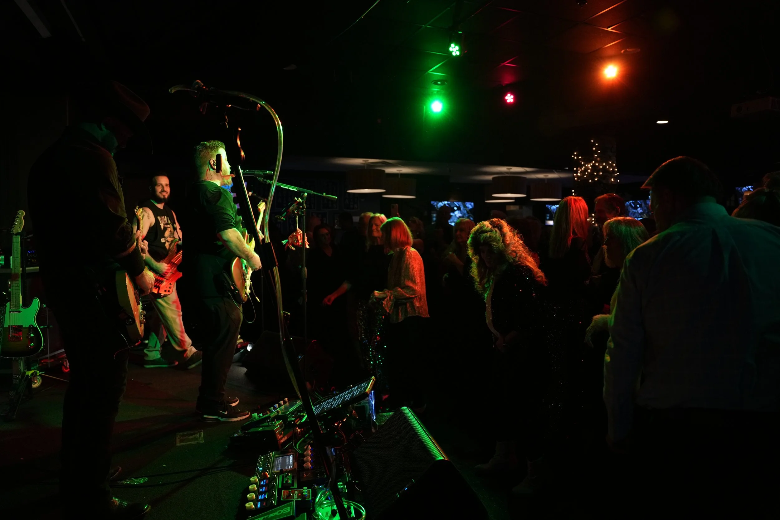 A live music event with a band performing on stage in front of an audience in a dark venue illuminated by green, red, and yellow stage lights. The band members are playing guitars and singing.