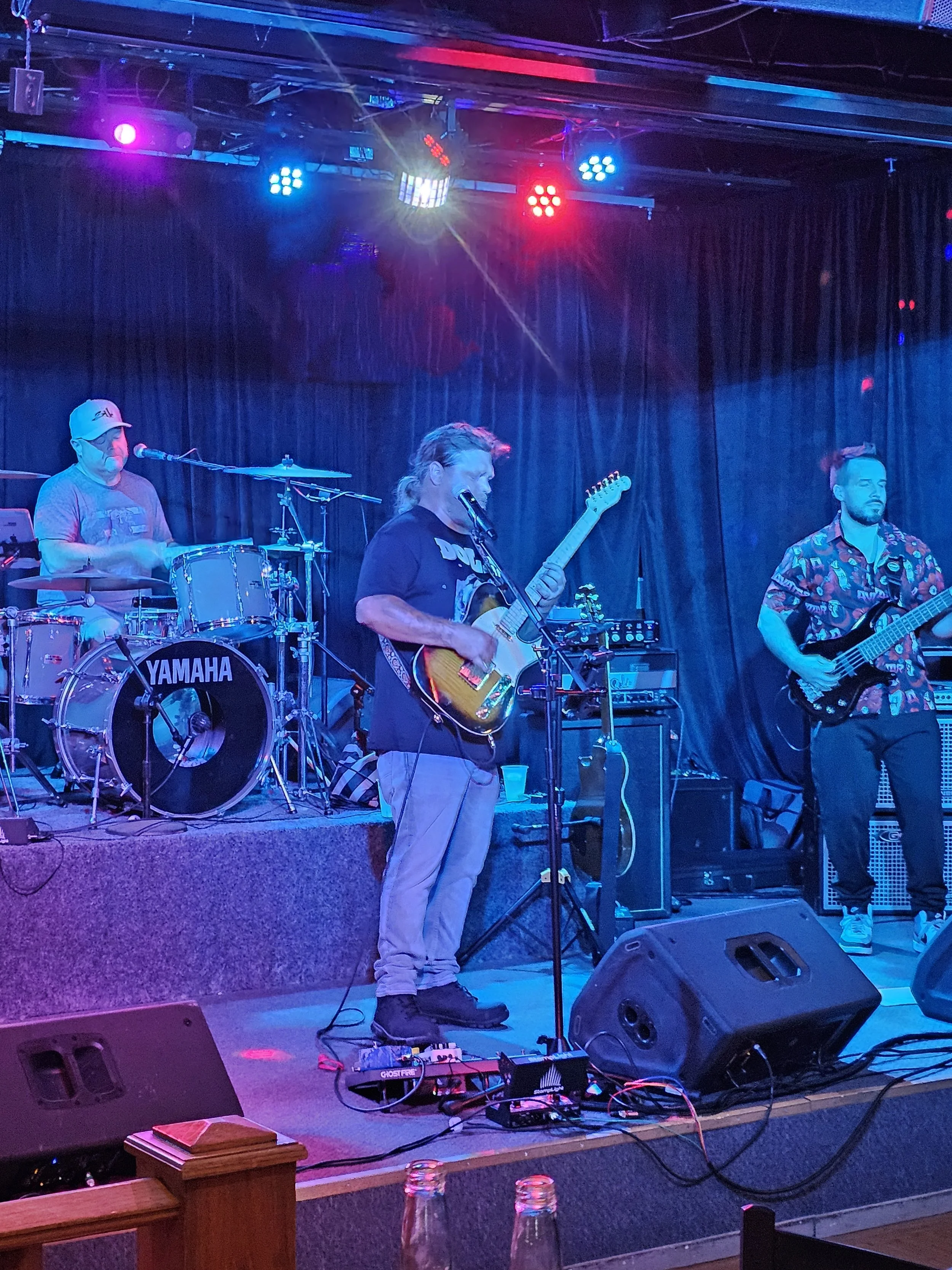 A live music performance on stage with three musicians playing instruments: one on drums, one on guitar, and one on bass. Stage is illuminated with colorful blue and red lighting. Black curtain backdrop. Foreground shows a small wooden railing and gl