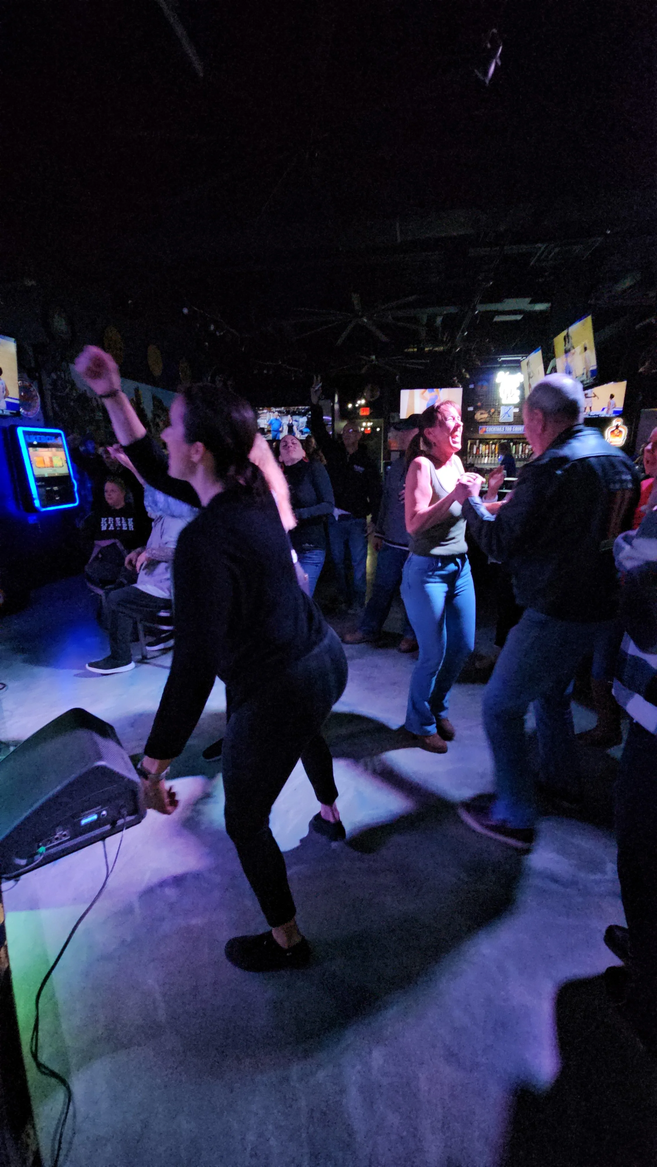 People dancing and socializing in a dark, lively bar or club with multiple TV screens on the wall.