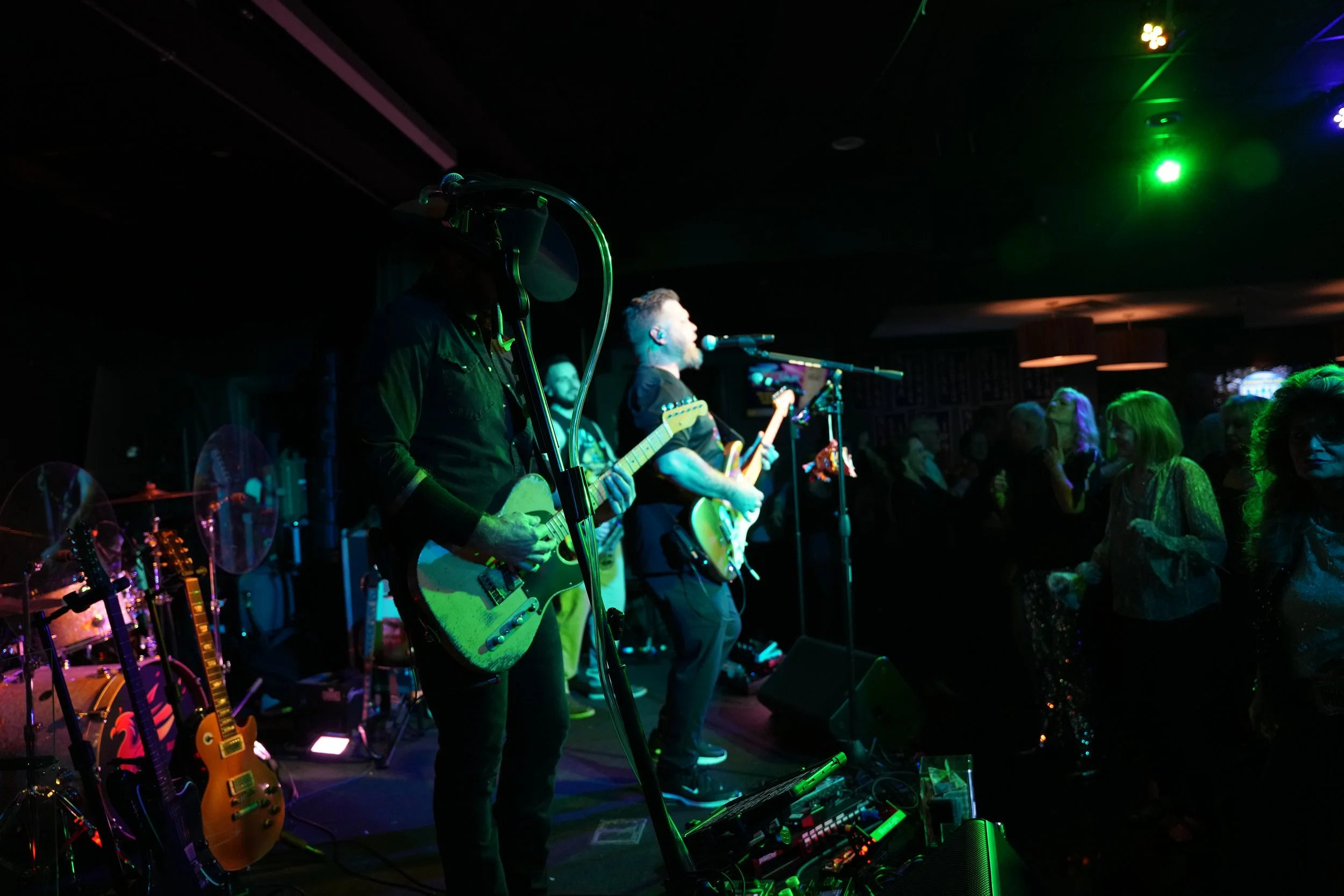 A band is performing on stage with guitars and microphones in a dimly lit venue, with audience members watching.