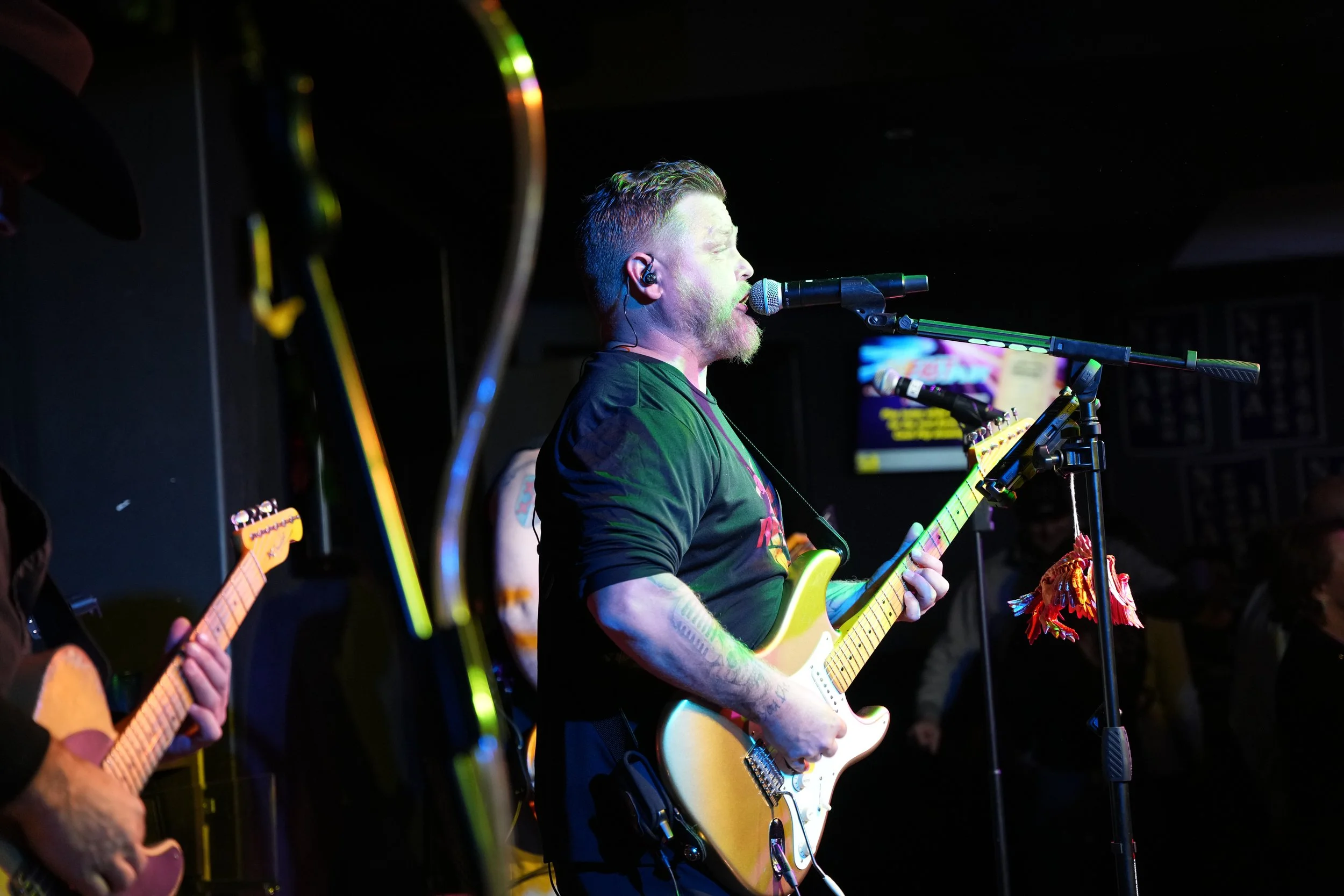A musician with short hair and tattoos playing an electric guitar on stage, singing into a microphone, with other guitar players partly visible in the background.