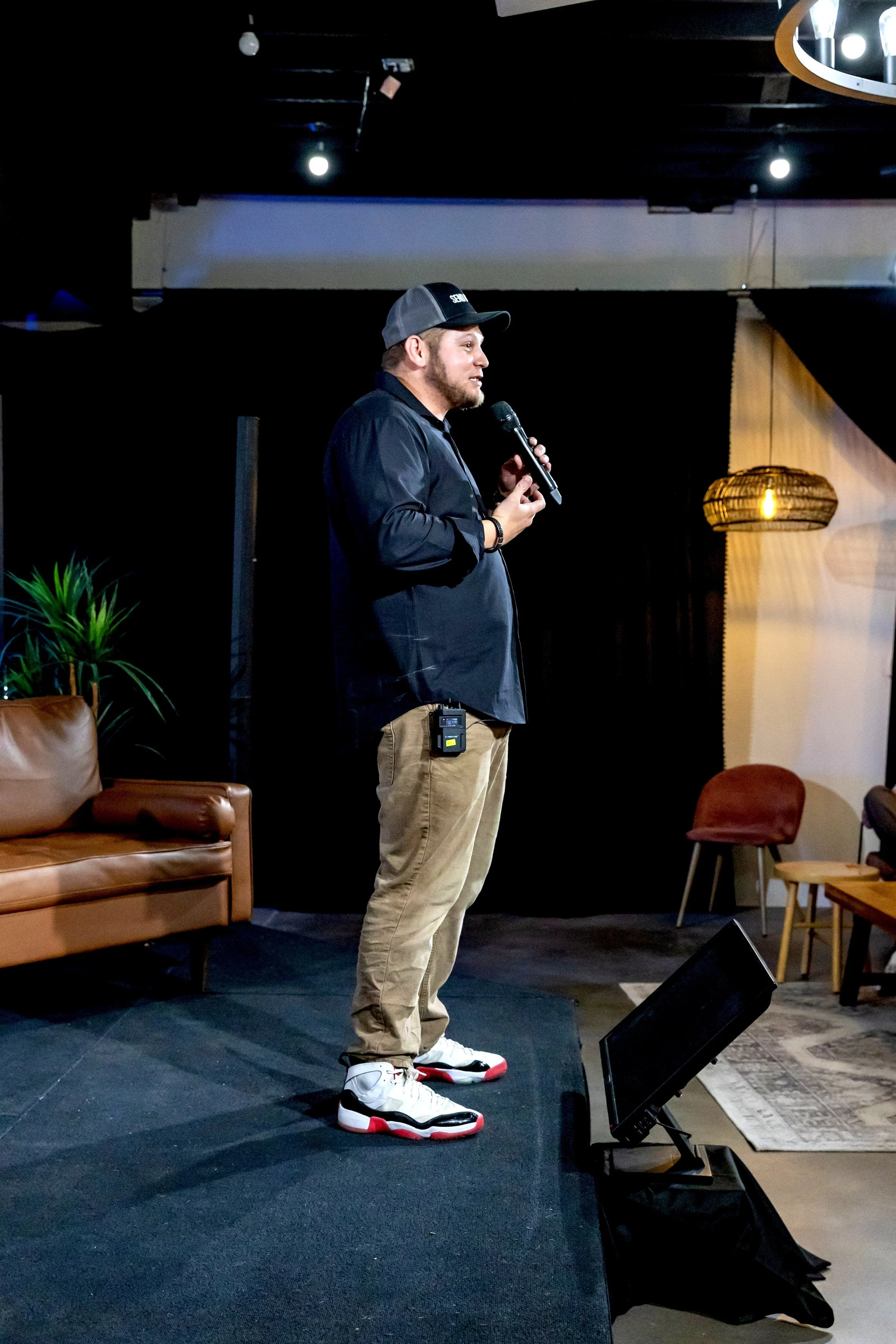 A man standing on a stage, holding a microphone and speaking to an audience. He is wearing a black cap, black shirt, khaki pants, and sneakers. There are chairs, a lamp, and a green plant in the background.
