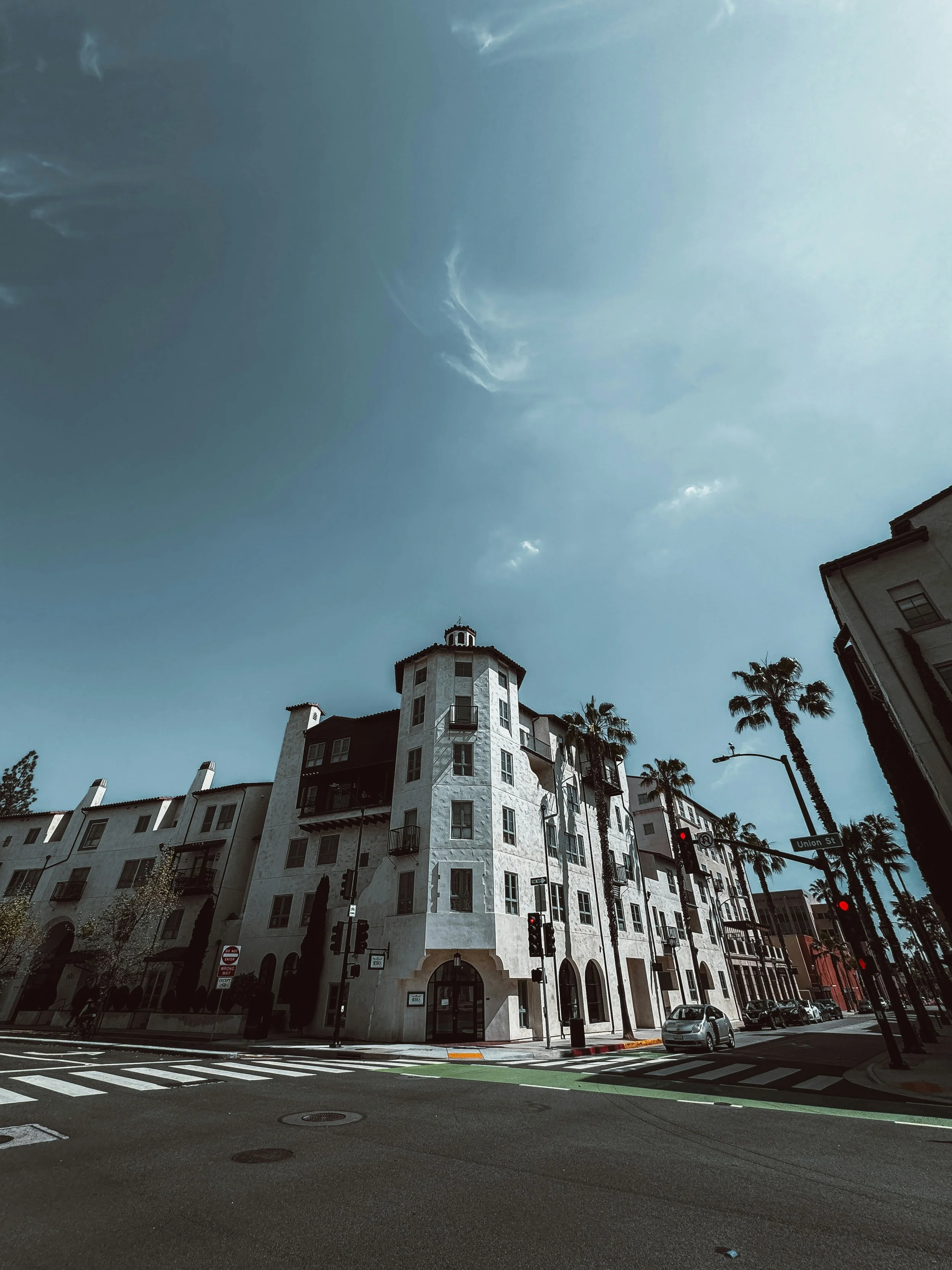 A city street scene featuring a large, white multi-story building with rounded towers on the corners, palm trees, cars parked along the curb, traffic lights, and street signs on a sunny day with clear skies.