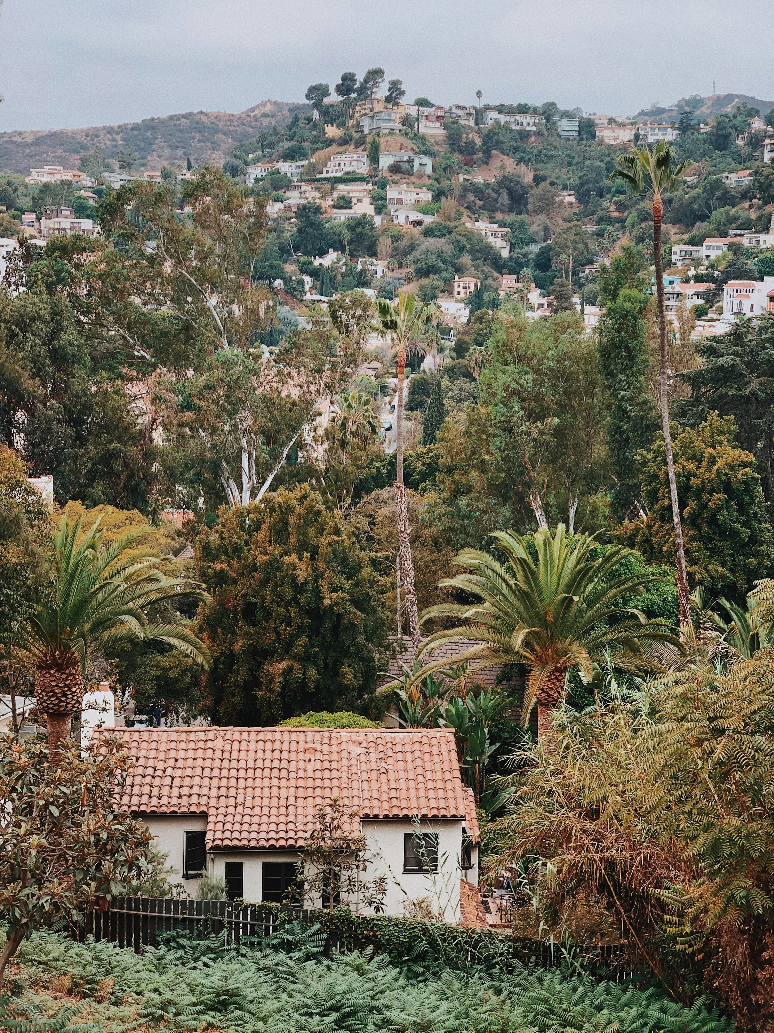 A house with a red-tiled roof surrounded by trees and foliage in a hilly landscape with numerous other houses in the background.