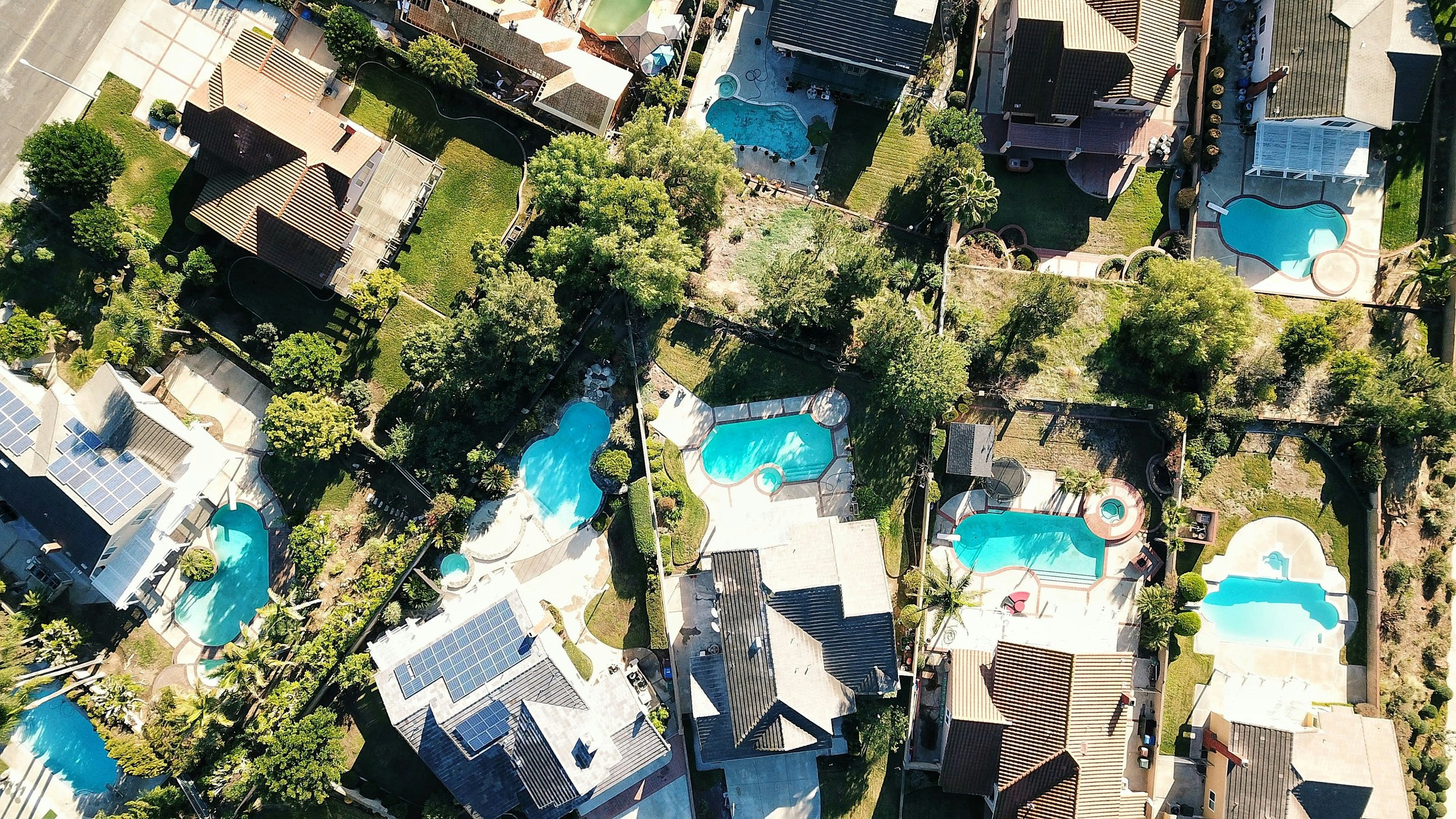 Aerial view of residential houses with swimming pools, trees, and solar panels on some rooftops in a neighborhood.