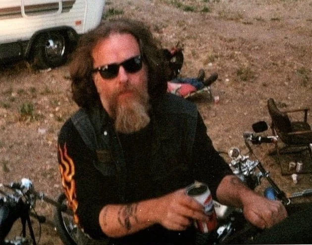 A man with long curly hair, a beard, sunglasses, and tattoos, holding a soda can, sitting outdoors near bicycles and camping gear.