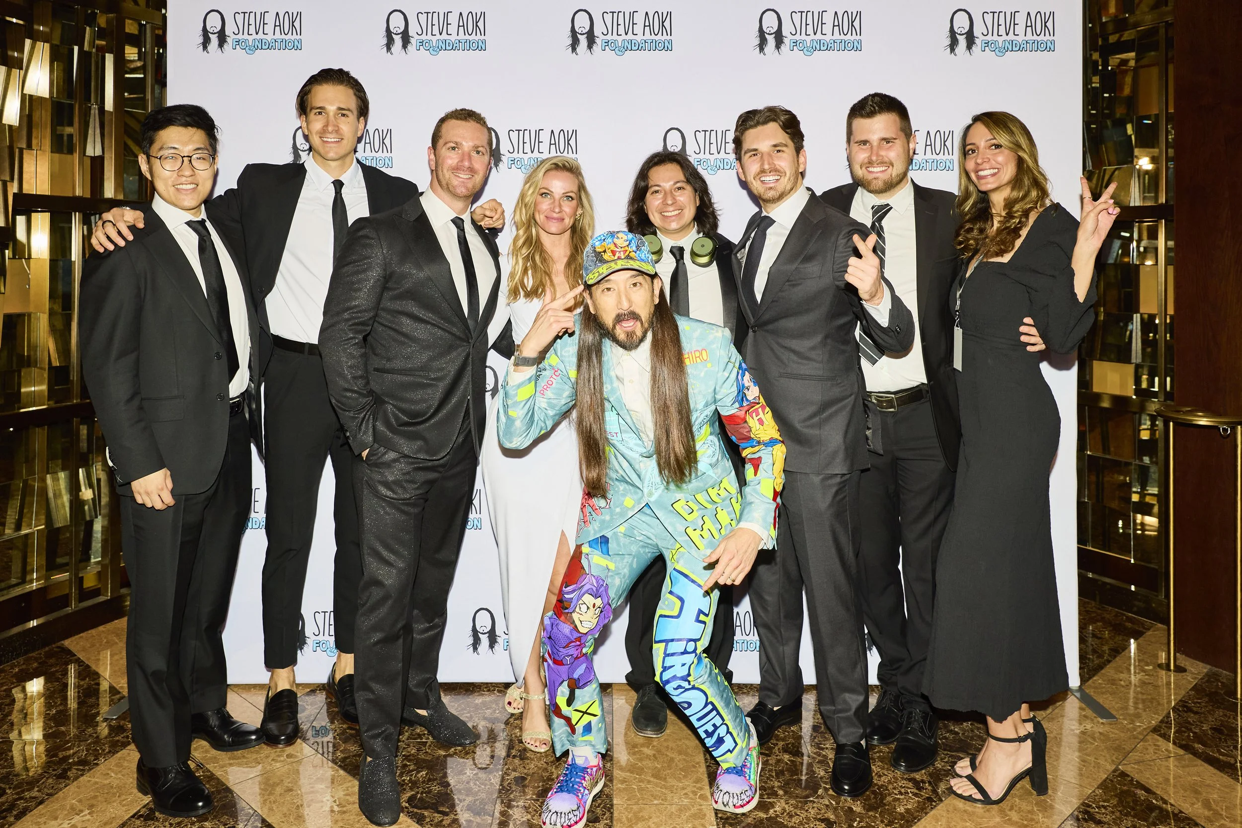Group of formally dressed guests pose on the red carpet at the 2025 Steve Aoki Foundation charity gala, standing in front of a branded step-and-repeat backdrop during the event.