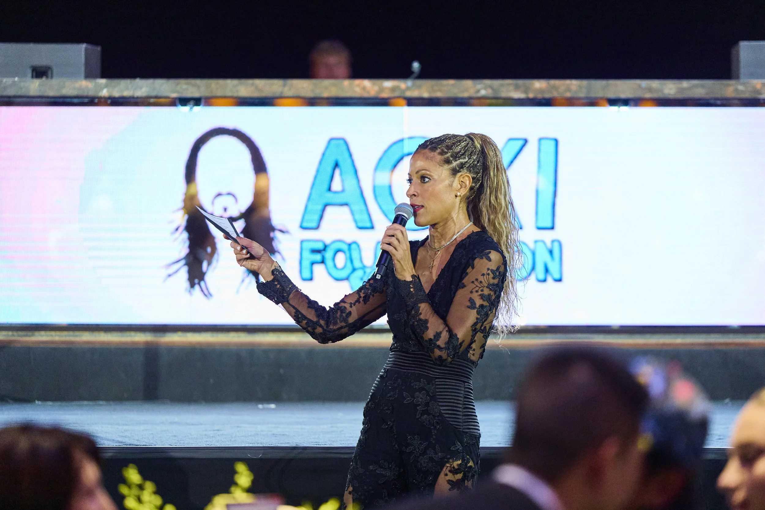 A professional auctioneer addresses the audience on stage at the 2025 Steve Aoki Foundation charity gala, holding a microphone and bid card during a live fundraising auction in a formal event setting.