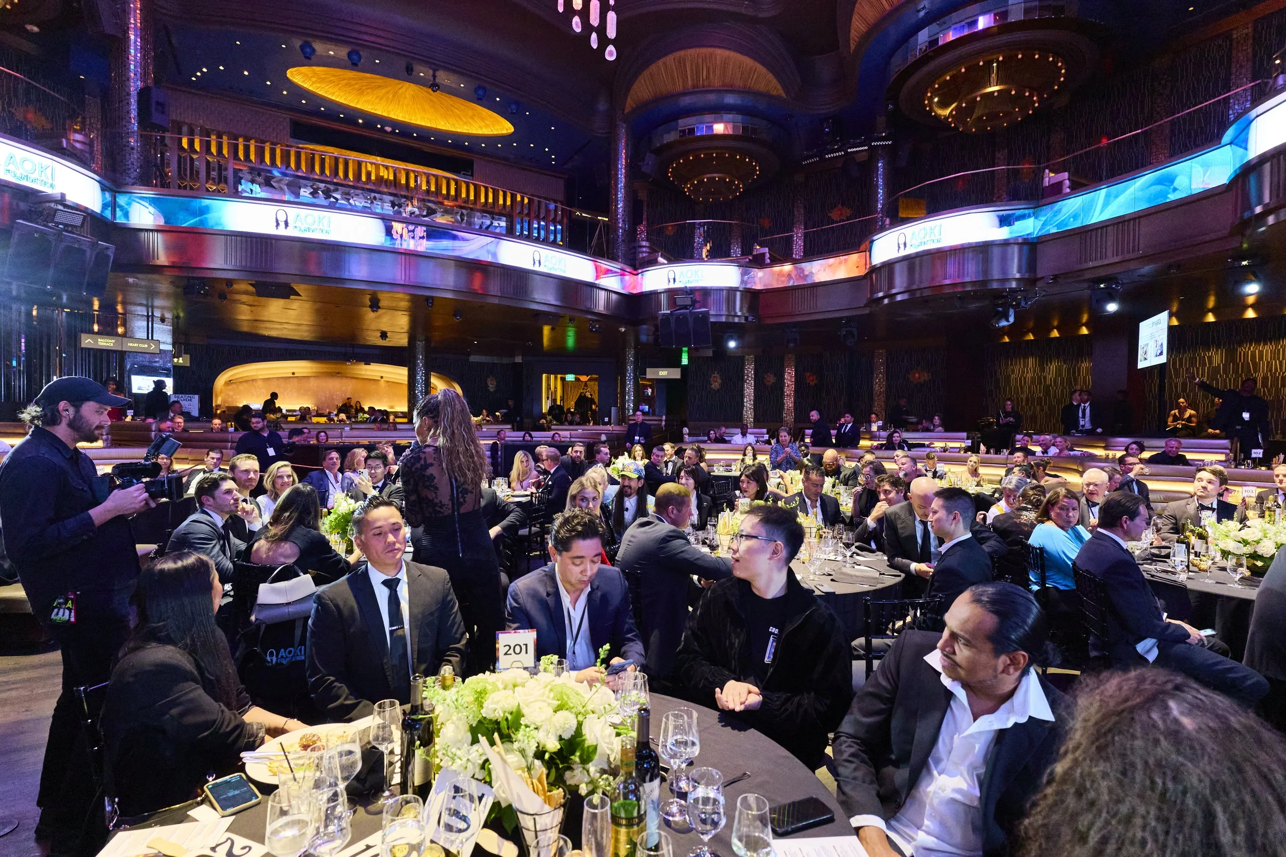 Wide-angle view of the 2025 Steve Aoki Foundation charity gala, featuring elegantly dressed guests seated at round tables inside a luxurious, multi-level venue during the live program.