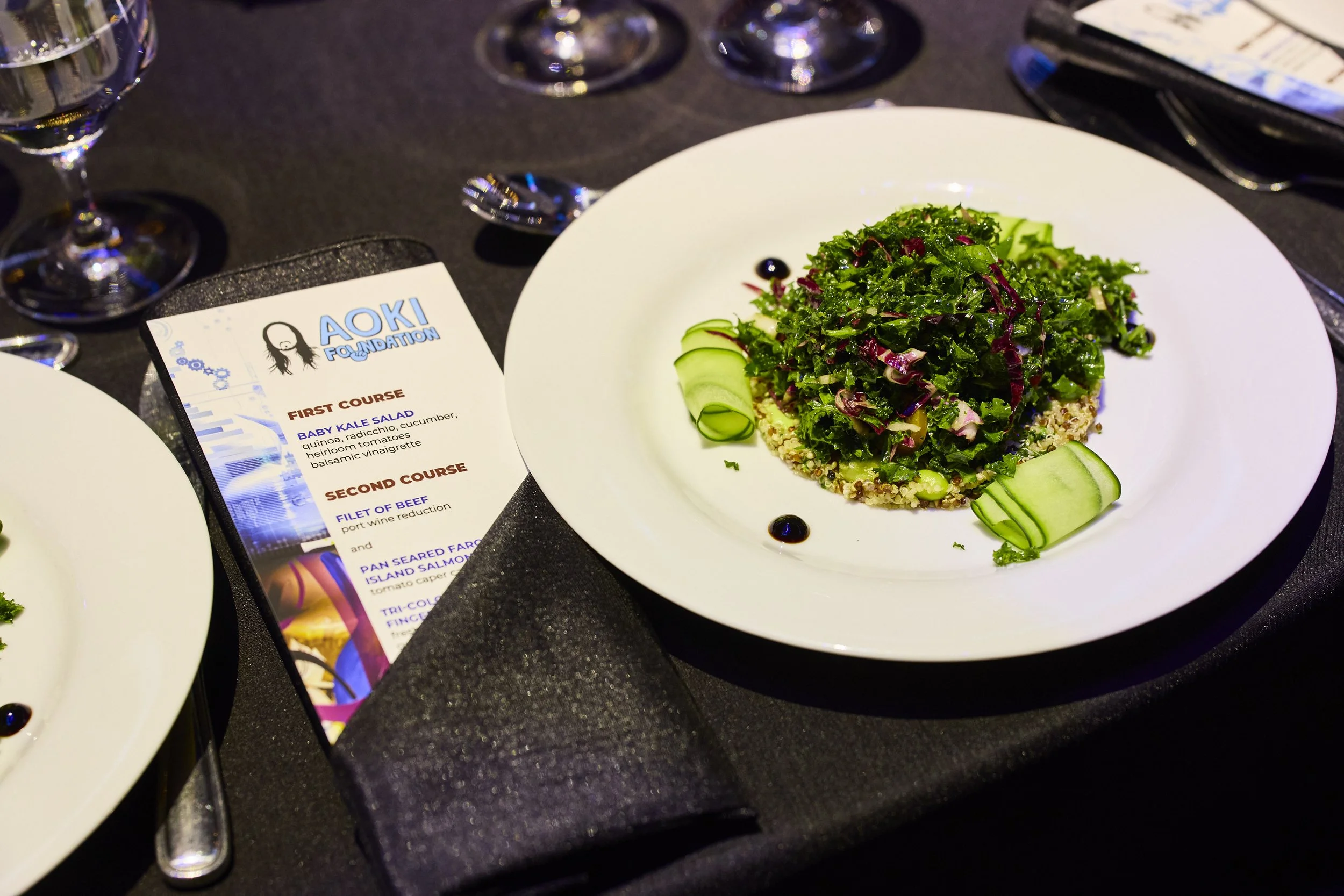 Plated gourmet kale salad served as a first course at the 2025 Steve Aoki Foundation charity gala, photographed alongside the printed event menu on a formally set table with wine glasses and linens.