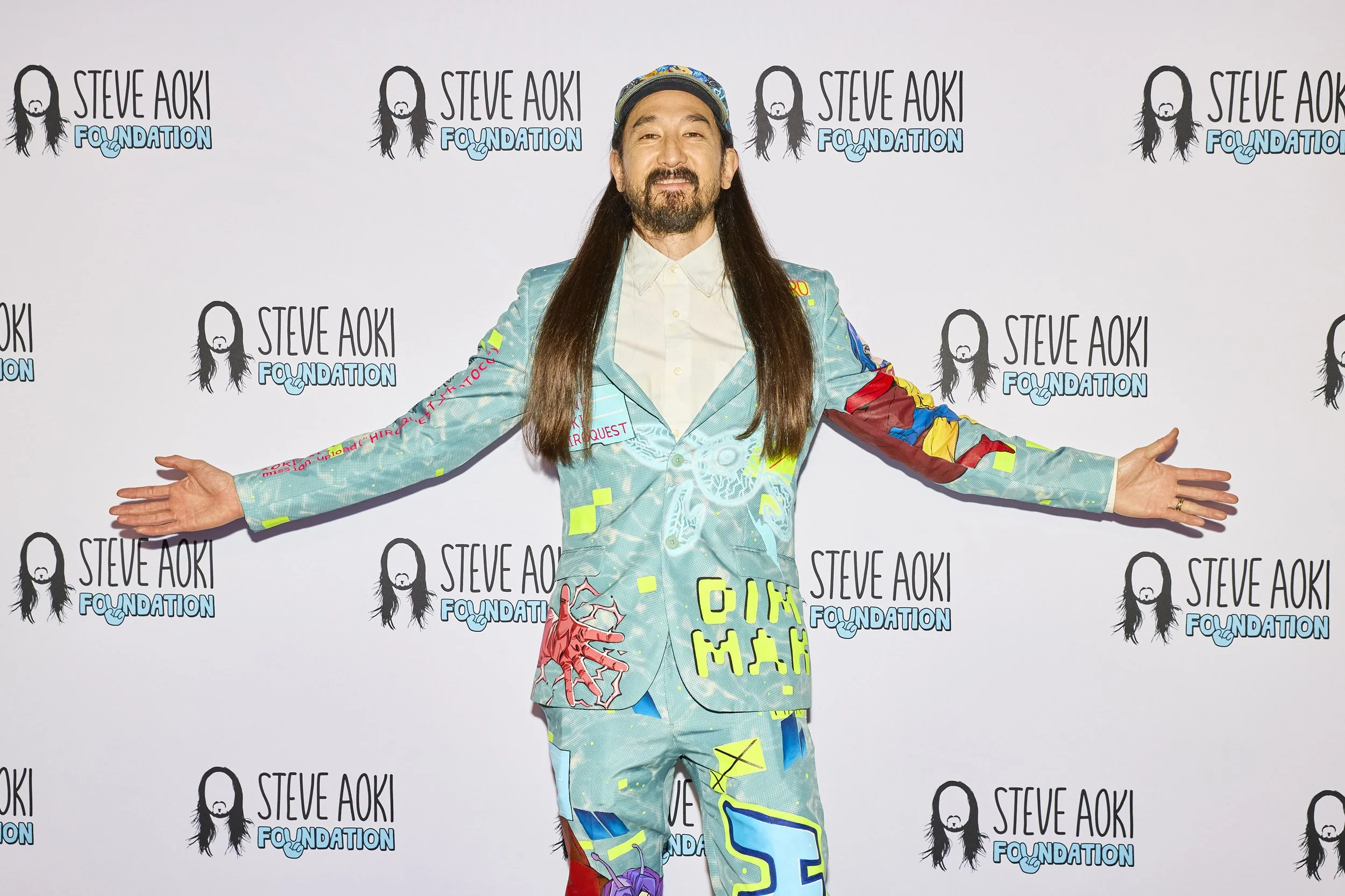 The event host poses with arms outstretched in front of a branded step-and-repeat backdrop at the 2025 Steve Aoki Foundation charity gala, wearing a colorful suit during the red carpet photo session.