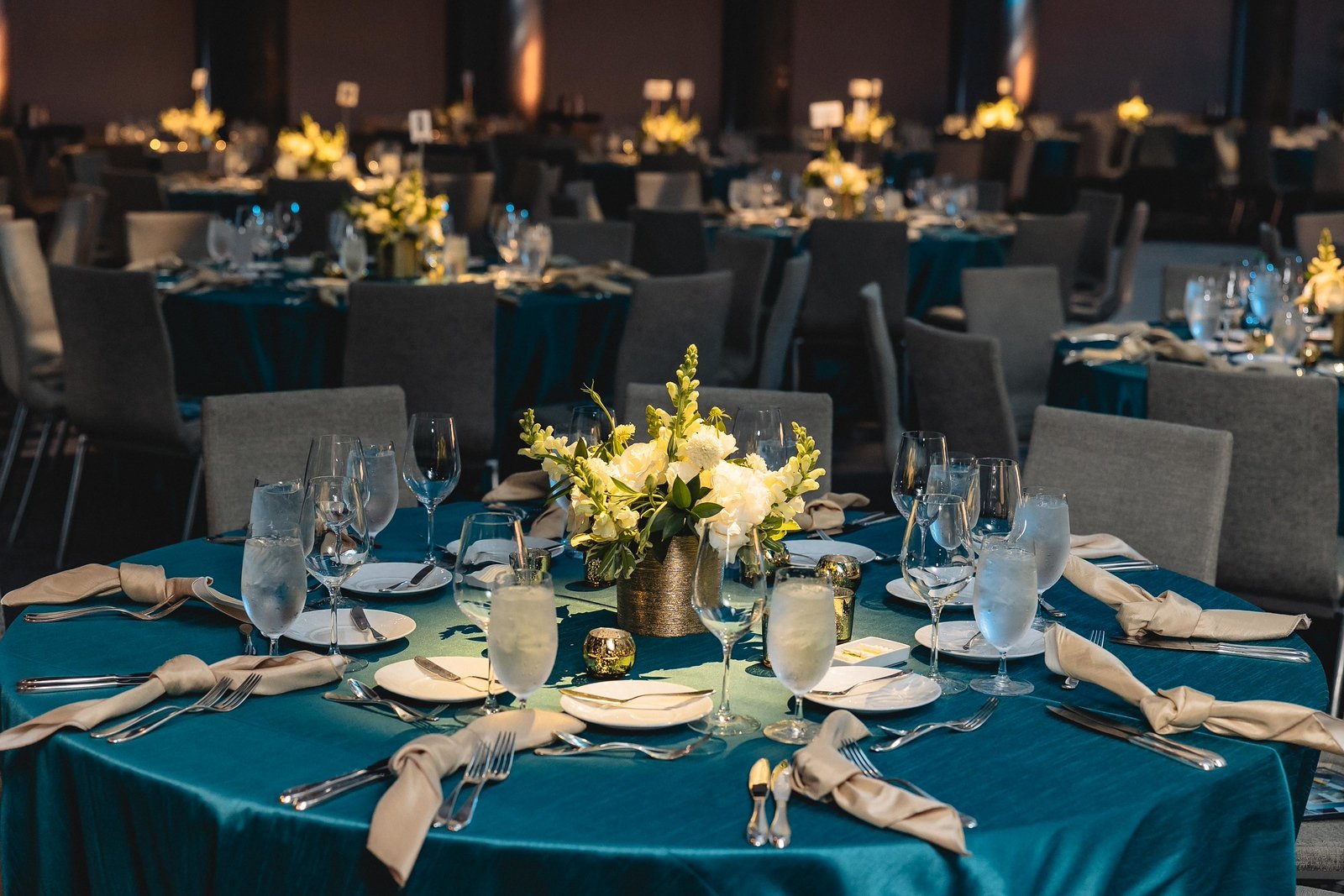 Up close shot of table settings at the Echo Horizon gala showcasing stunning white floral arrangements, white and silver cutlery/glasses/plates, dramatic lighting, and blue satin table coverings. 