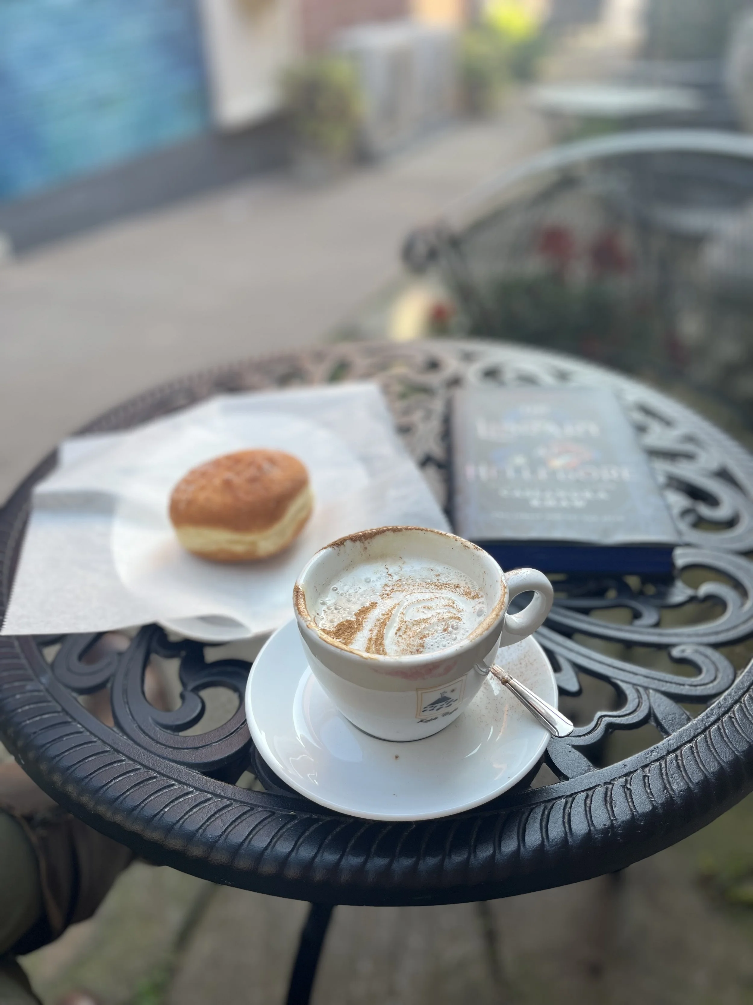 A delicious donut, coffee, and book in focus on a wrought iron table