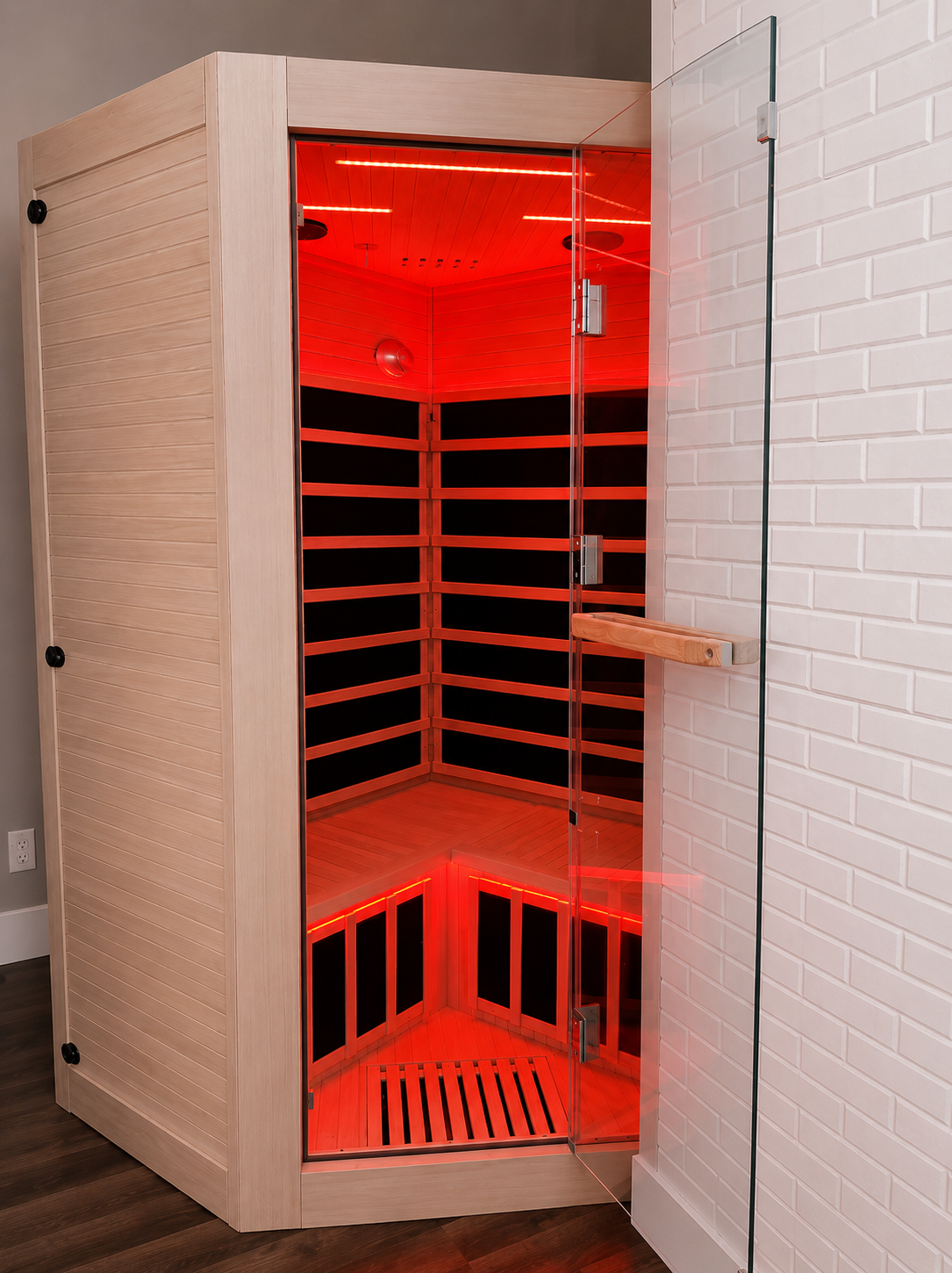 A modern infrared sauna with wooden interior and black panels, enclosed by glass door, located next to a white brick wall.