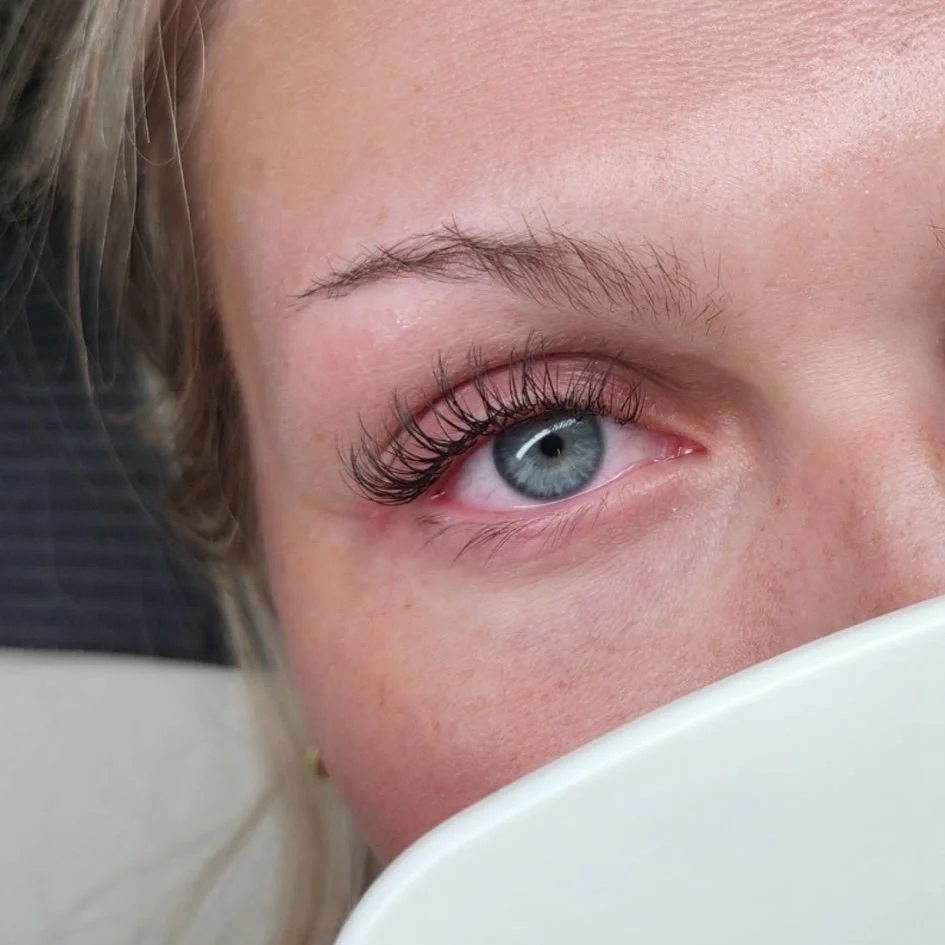 Close-up of a person's face focusing on their blue eye, with long eyelashes and well-groomed eyebrows, partially obscured by a white object.