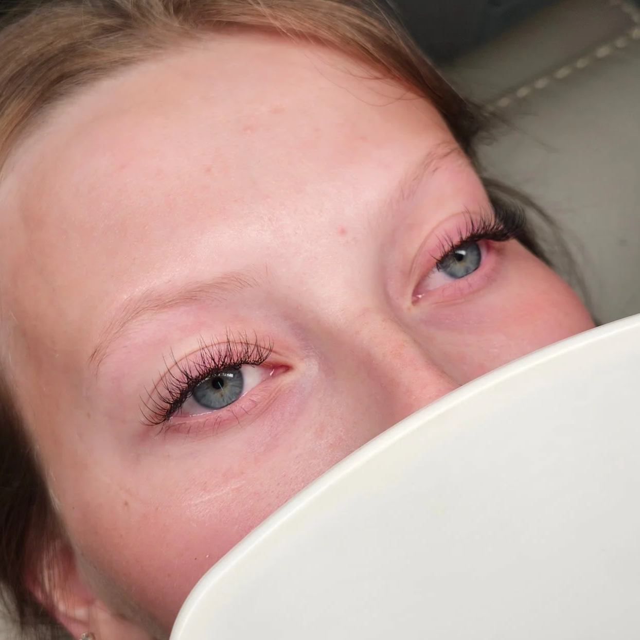 Close-up of a woman with light skin and blue eyes, lying down with her face partially covered by a white object, showing her facial features and makeup on her eyelashes.