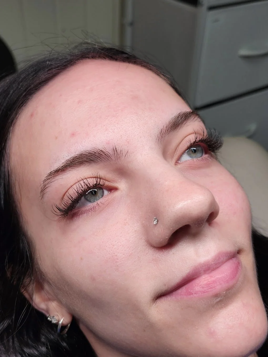 Close-up of a young woman's face with long, curled eyelashes, a nose piercing, and earrings, lying back with her eyes looking upward.