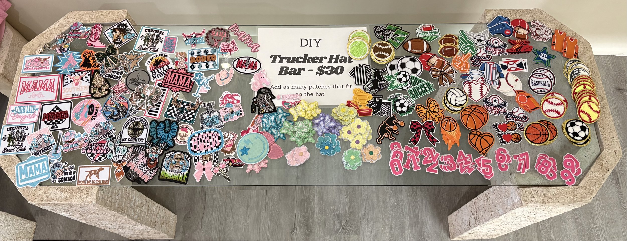 Table displaying various patches and stickers for a DIY Trucker Hat Bar, including sports, sports teams, animals, and decorative patches, with a central sign reading 'DIY Trucker Hat Bar - $30'.