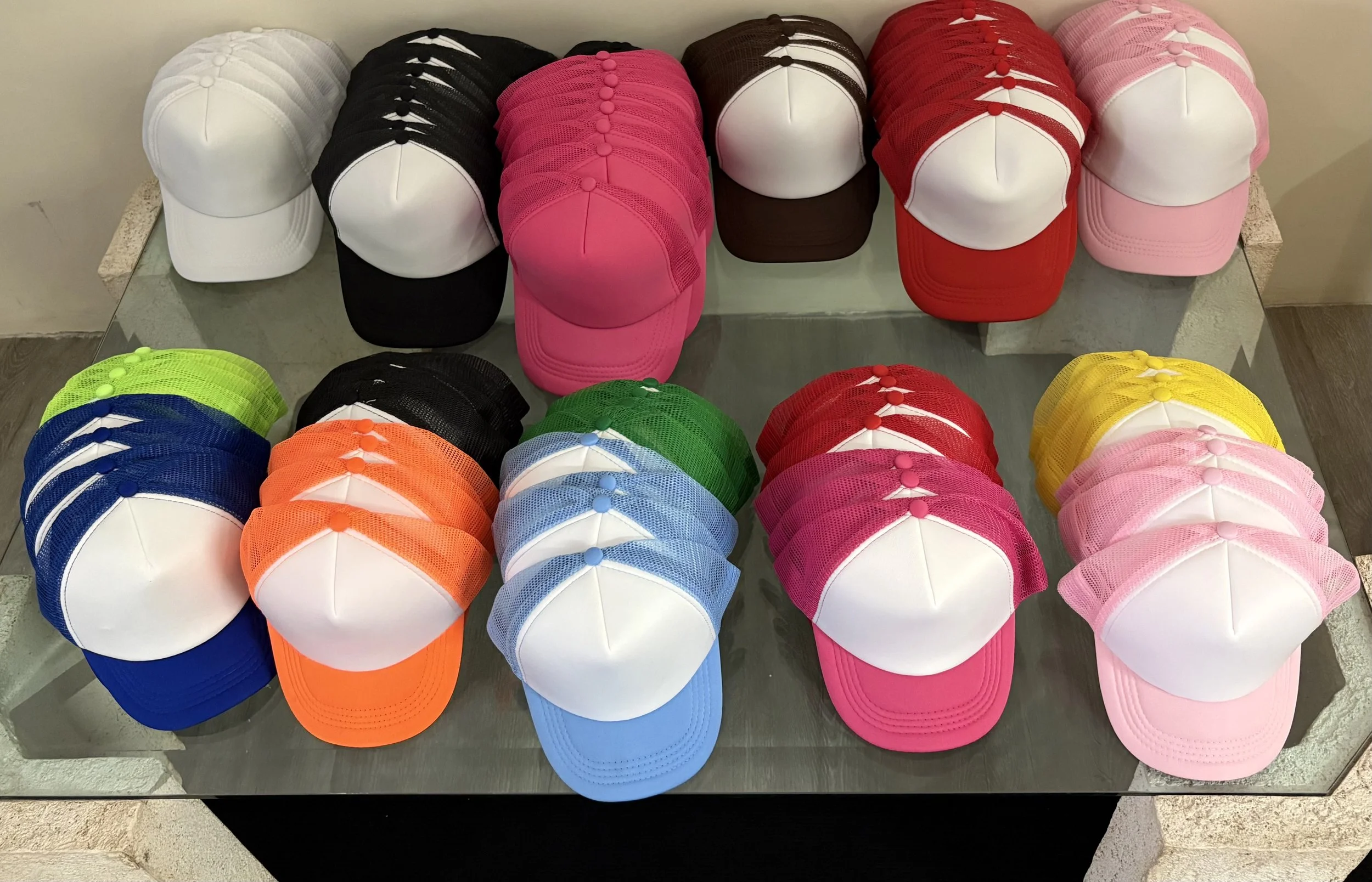 Multiple colorful baseball caps displayed on a glass table, including white, black, pink, red, brown, blue, orange, green, yellow, and pink caps.