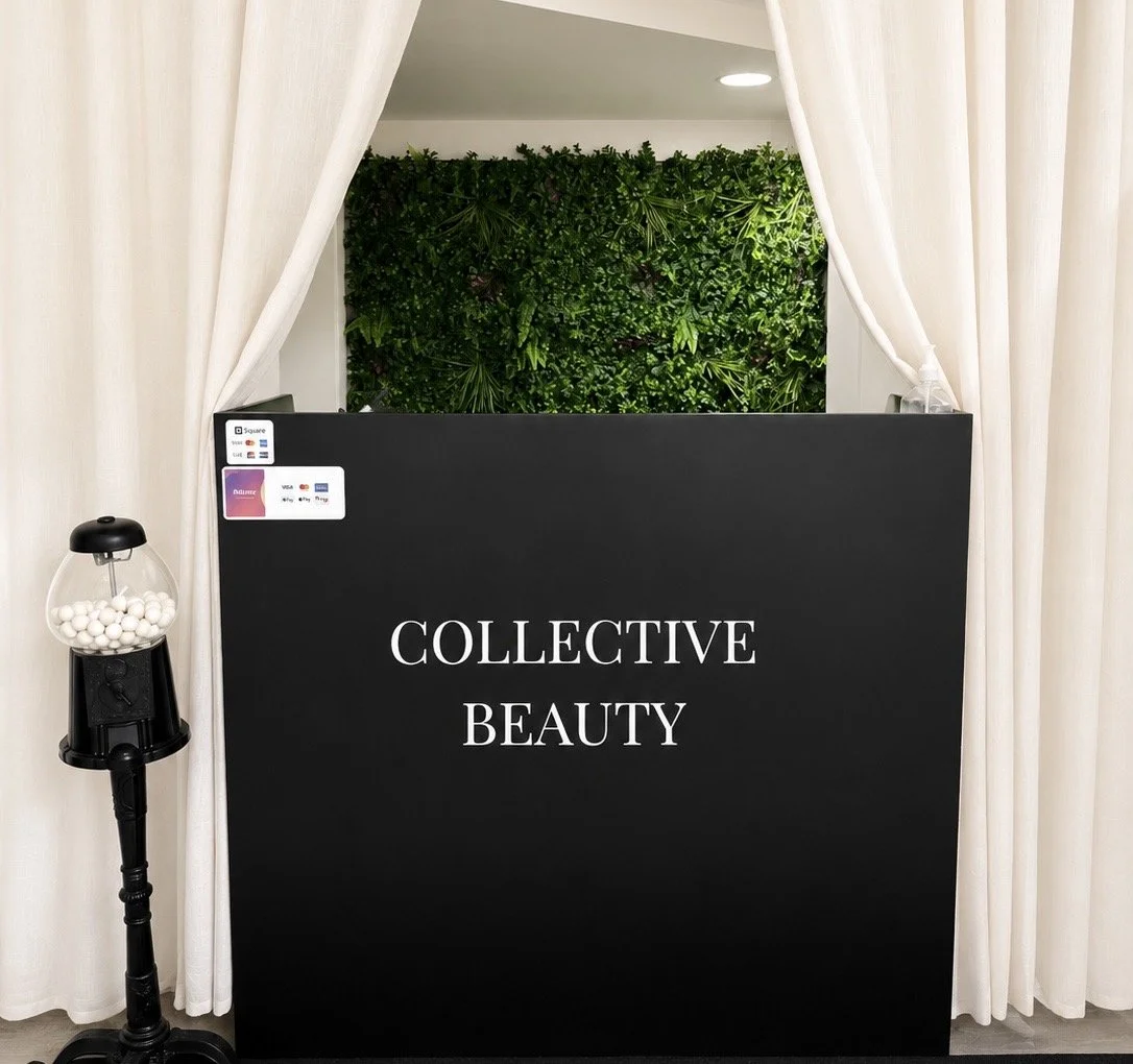 Reception desk with a black sign that says 'Collective Beauty,' white curtains on either side, a green plant wall behind, and a gumball machine to the left.