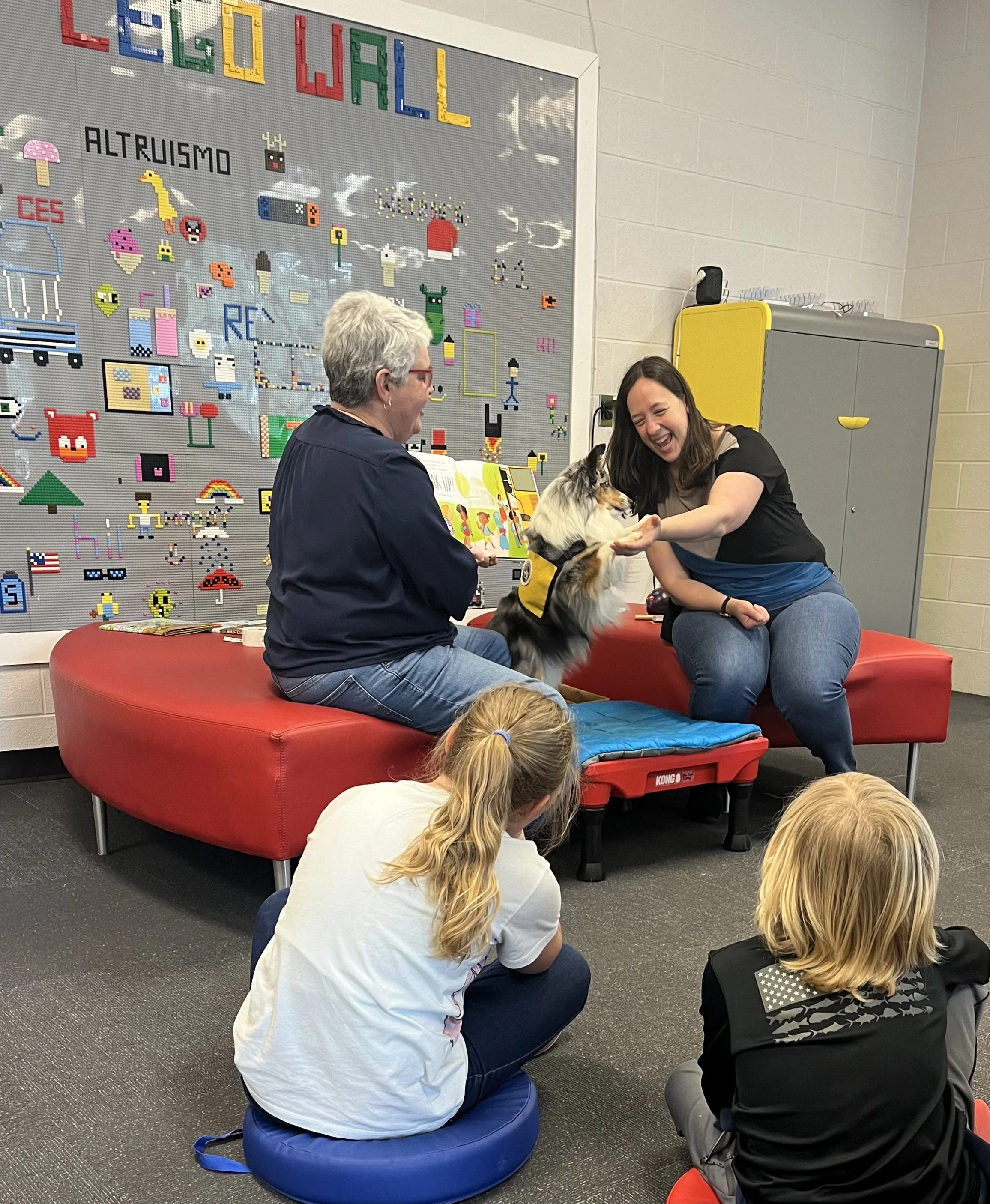 A therapy dog session with a dog sitting on a platform, interacting with a woman, while another woman reads aloud, and children watch. The setting is a classroom or library with a pixel art wall.