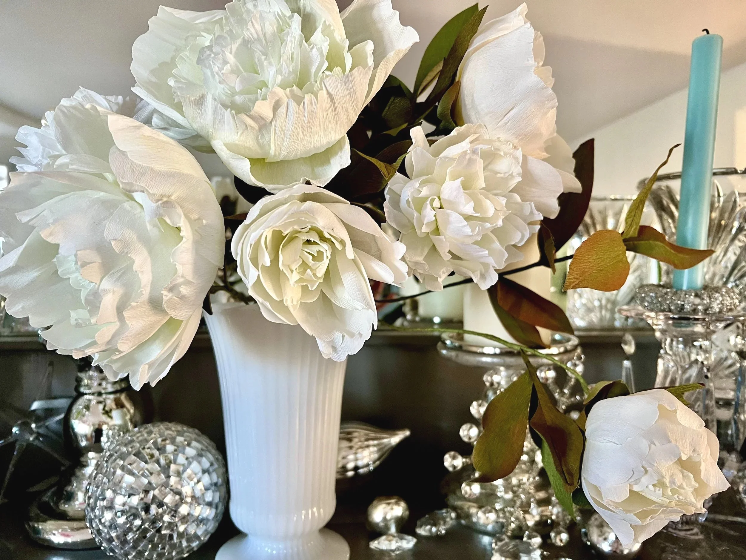 White peony flowers in a white vase surrounded by decorative glass and silver ornaments.