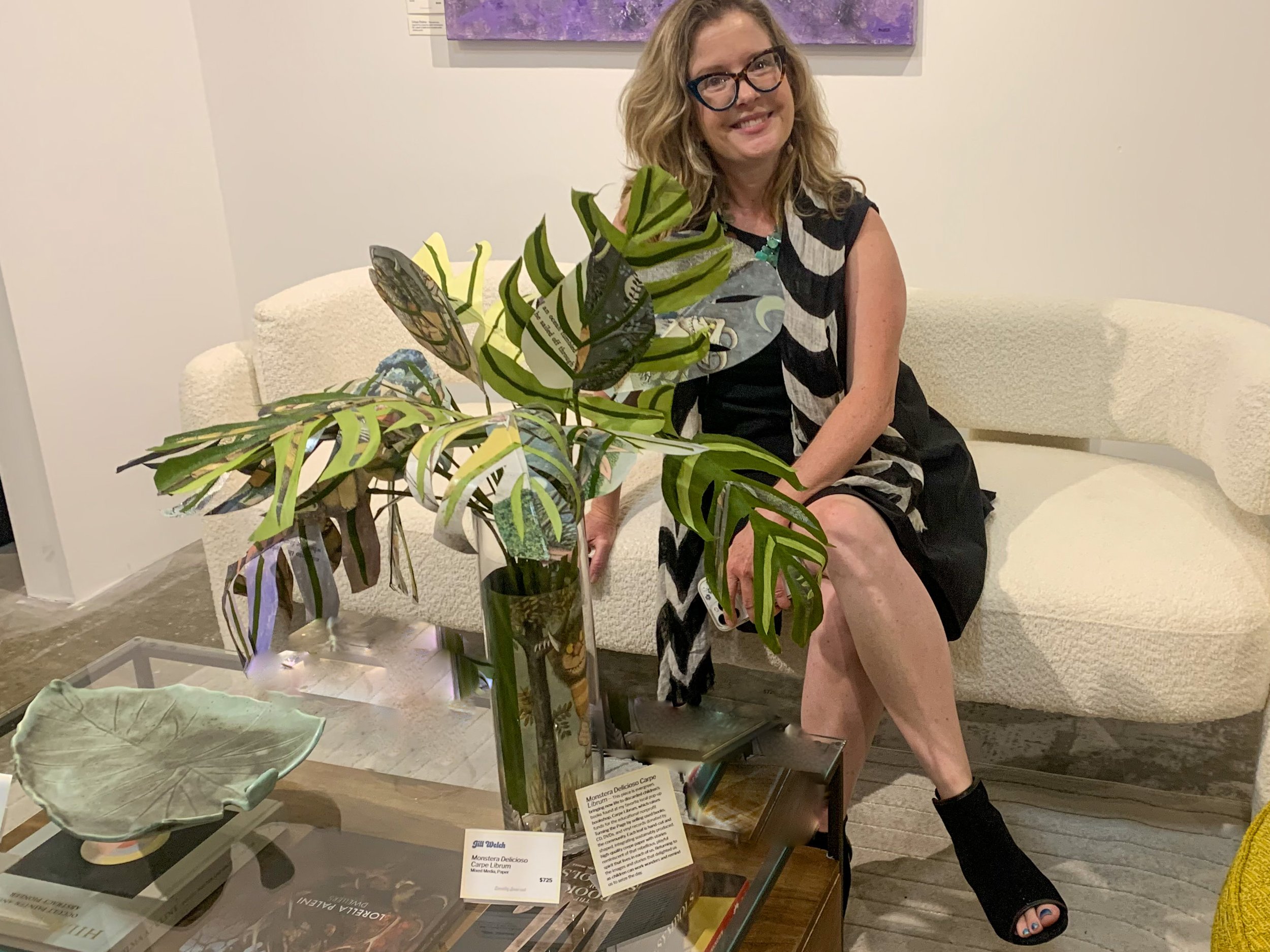 A woman sitting on a cream-colored sofa, wearing glasses and a black dress with a black and white patterned scarf, smiling at the camera. She has blonde, wavy hair and is holding a large green-leaved paper plant on a table in front of her.