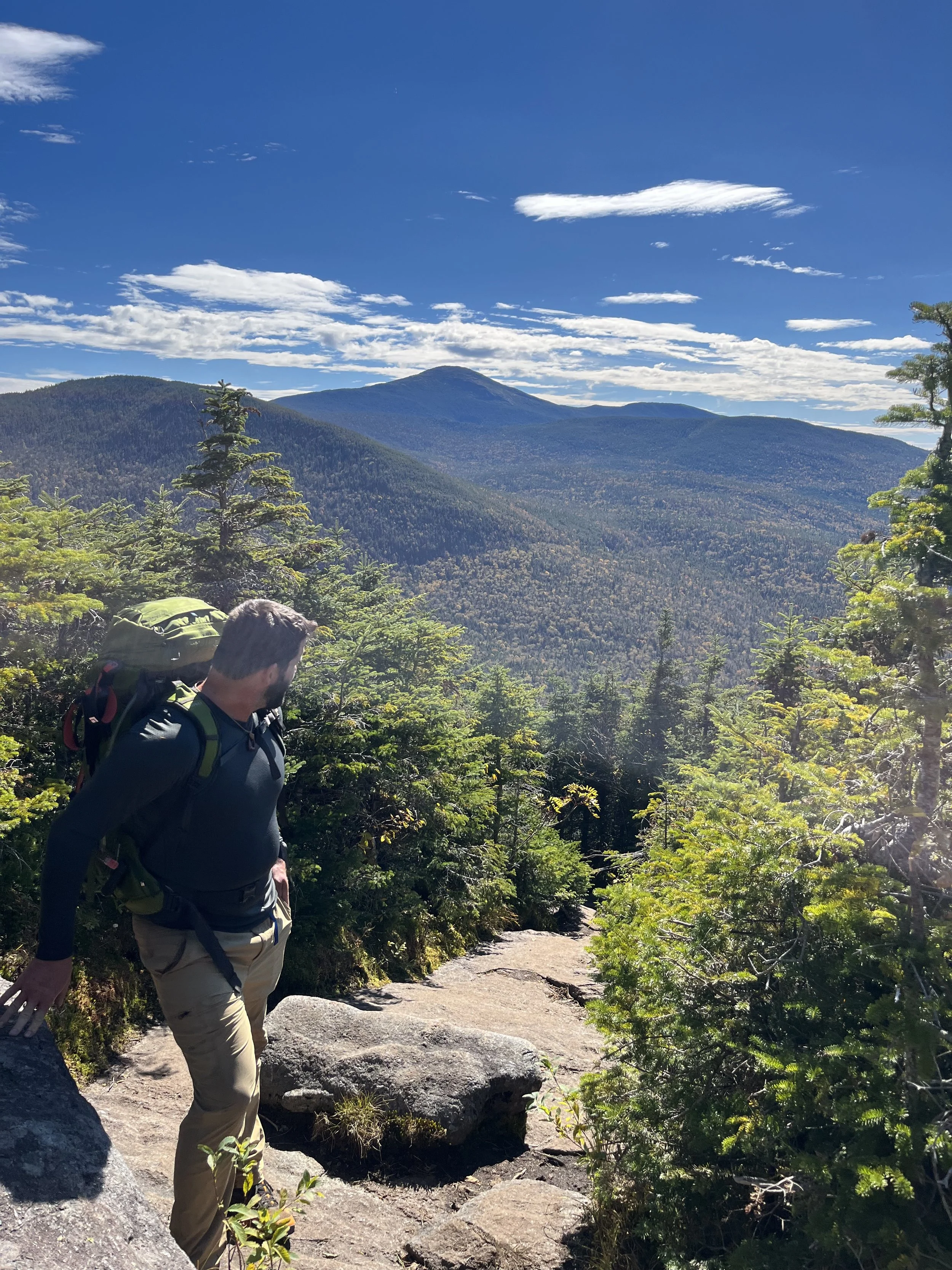 A man hiking on a mountain trail with a view of lush green trees, distant mountains, and a partly cloudy blue sky.