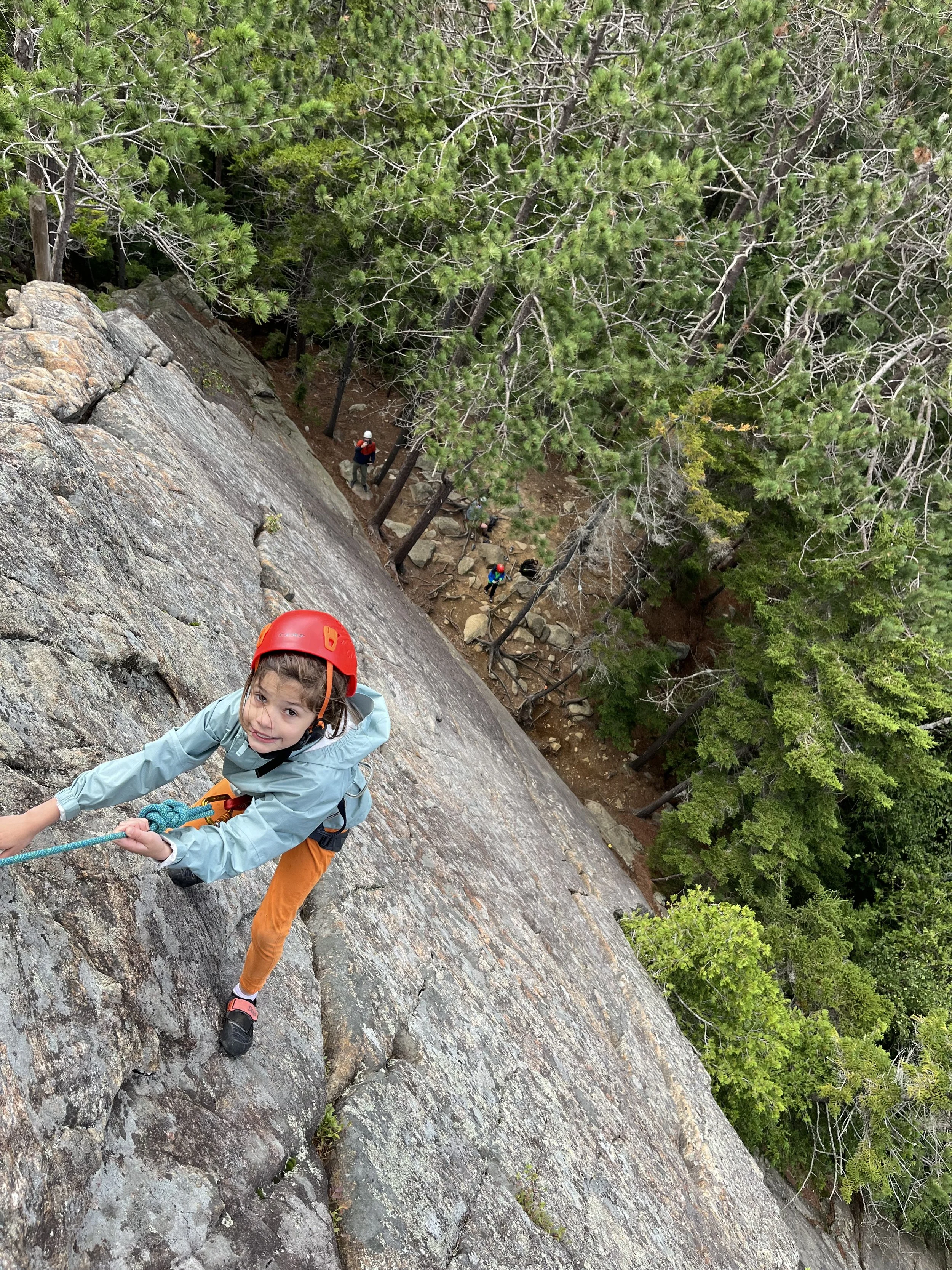 A young girl rock climbing on a steep, rocky surface with a red helmet and a light jacket, holding a climbing rope. In the background, there are additional climbers and a dense forested area with tall green trees.