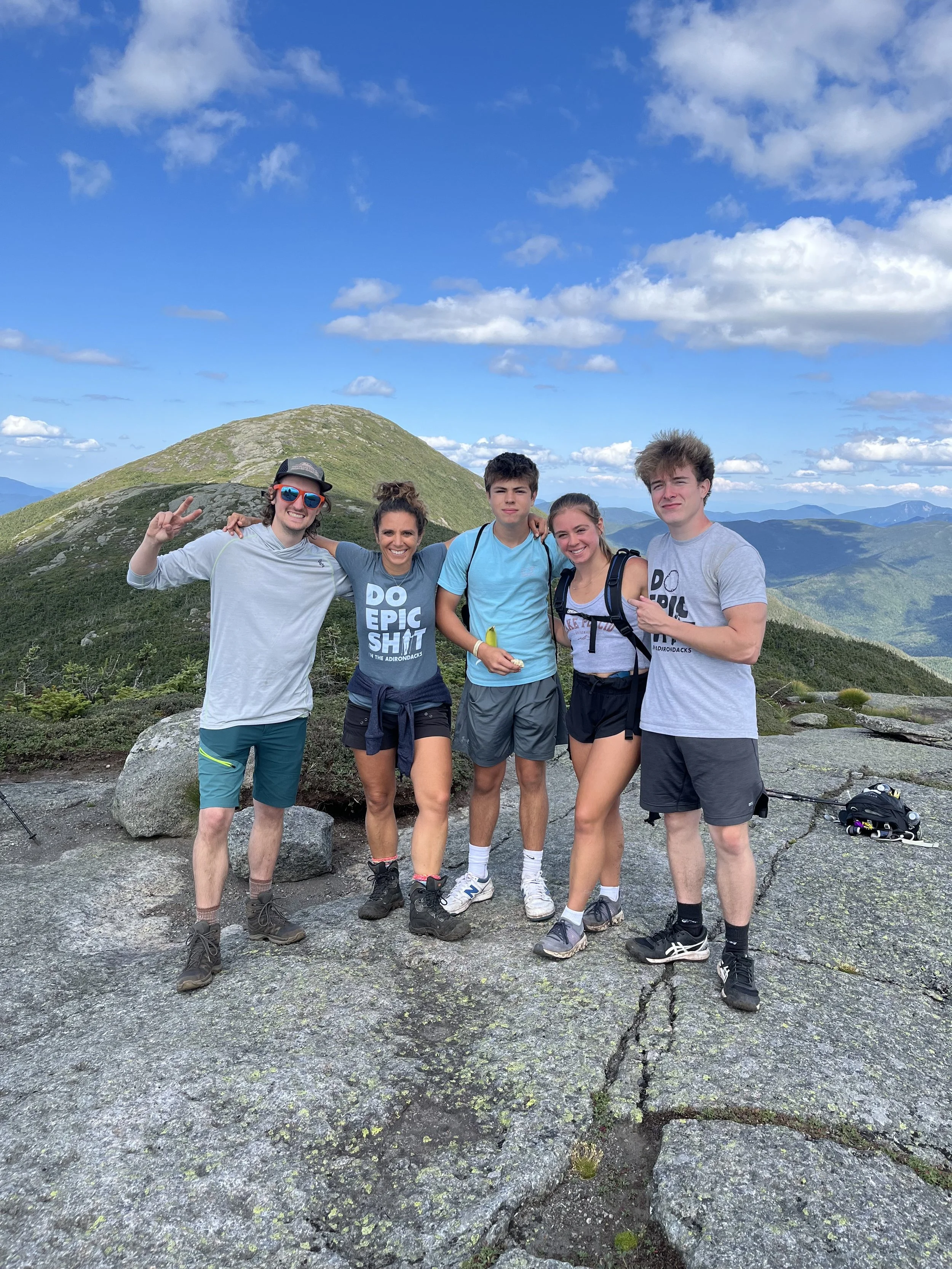 Group of five young people smiling and posing on a mountain trail with green hills and a partly cloudy sky in the background.