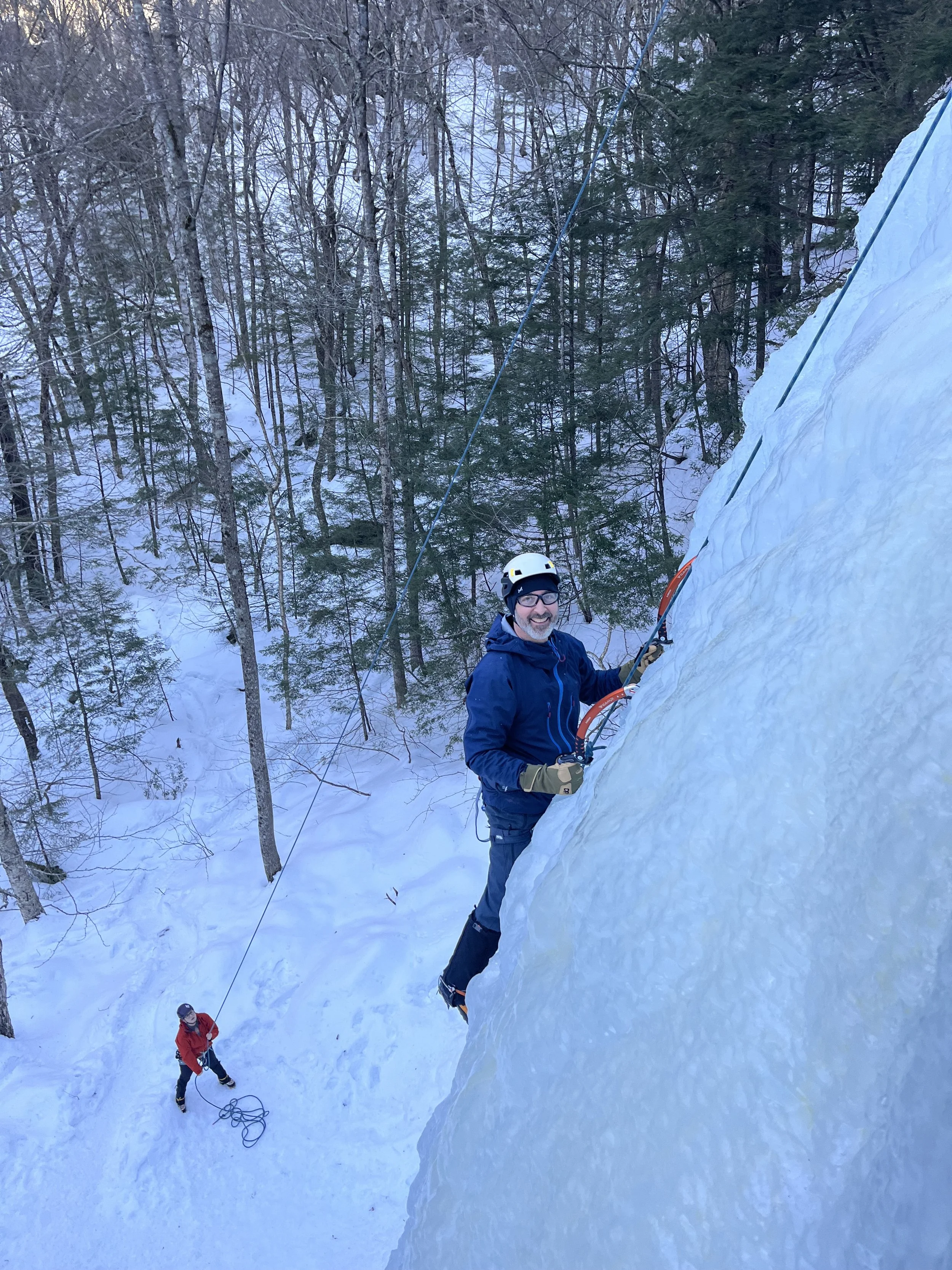 A man wearing a helmet and winter clothing is ice climbing on a frozen ice wall, using ice axes and crampons, while another person stands on the snowy ground holding a safety rope in a forested winter scene.