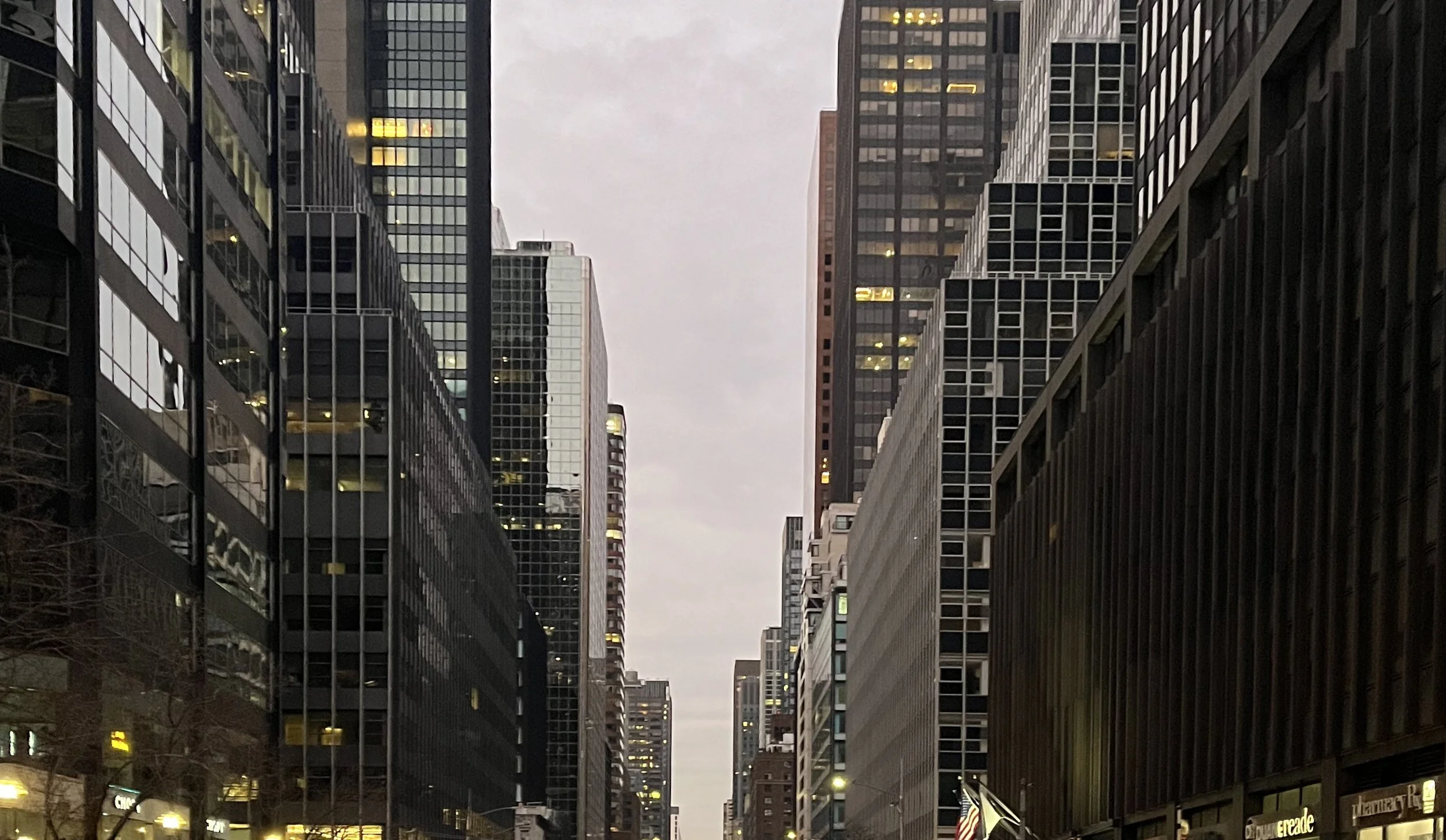A city street at dusk with tall modern glass office buildings on both sides, some windows illuminated, and an American flag on a pole near the street.