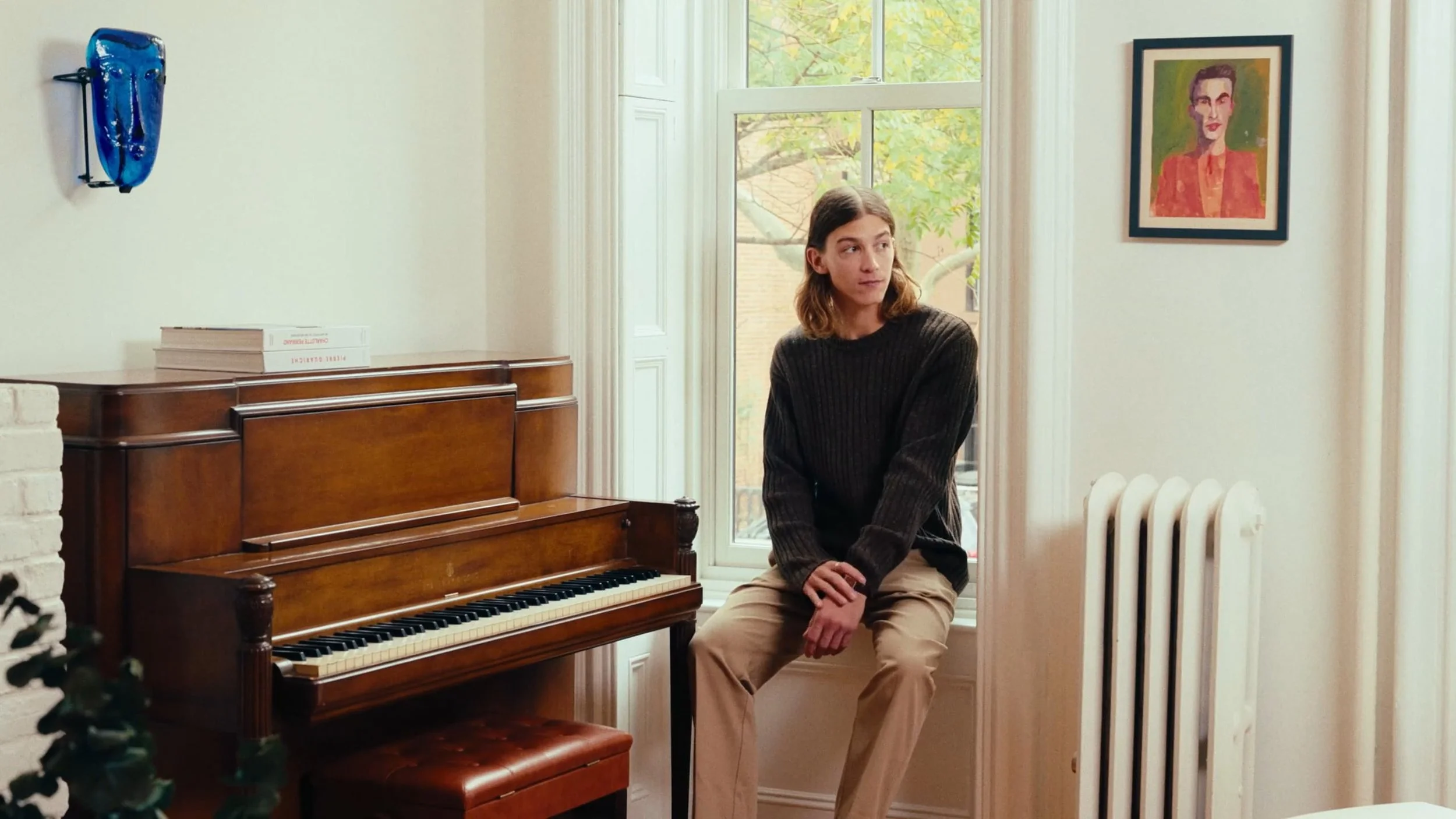 A young woman with long hair sitting on a windowsill next to a wooden upright piano in a bright room with white walls, a framed portrait of a woman on the wall, and a radiator behind her. Outside the window, trees with green leaves are visible.
