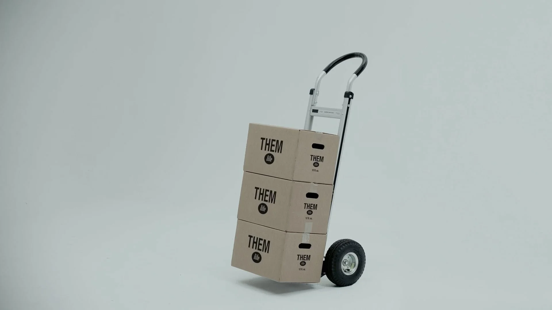 A hand truck loaded with three stacked cardboard boxes labeled 'THEM lite' and '12 oz.' against a plain light gray background.