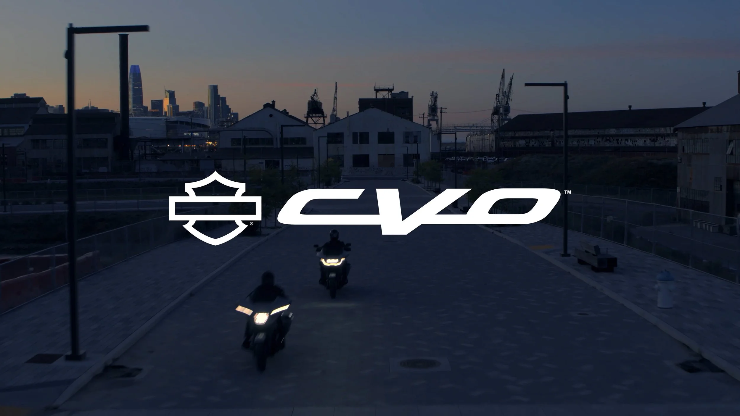 Two motorcyclists riding on a paved urban pathway during dusk with city buildings and cranes in the background, overlaid with the Harley-Davidson and EVOC logos.